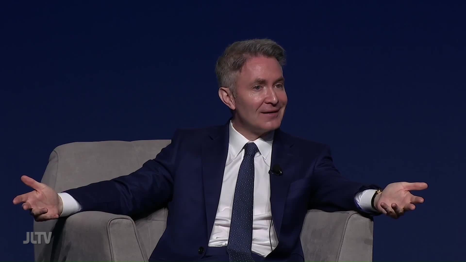 The man, seated in a chair, gestures expansively with both hands. He's on Jewish Life Television, speaking with a smile.
