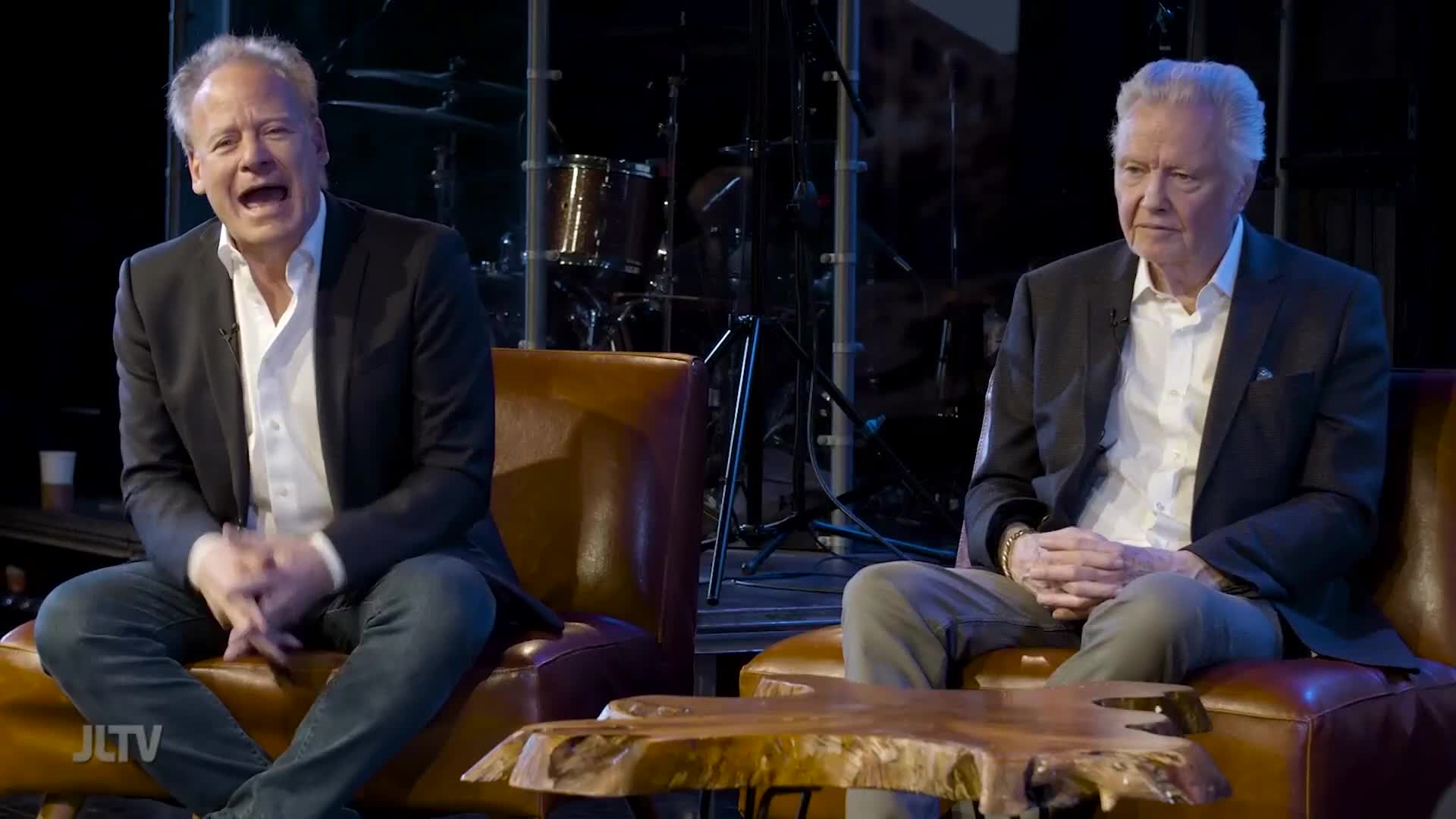 On a set for Jewish Life Television in the United States, two men sit in armchairs. The man on the left is animated, speaking with his hands, while the other listens intently.
