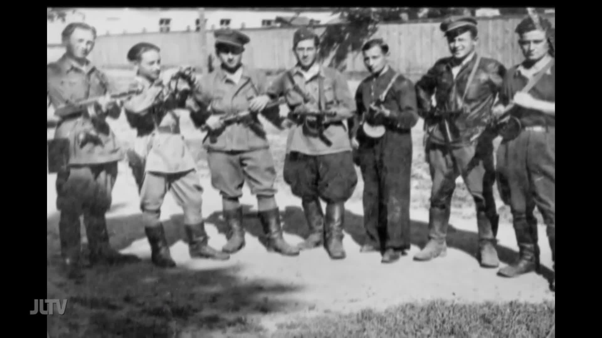 Eight men, all in military-style uniforms, stand shoulder-to-shoulder, each holding a rifle. The scene, broadcast by Jewish Life Television, is a stark black and white image.
