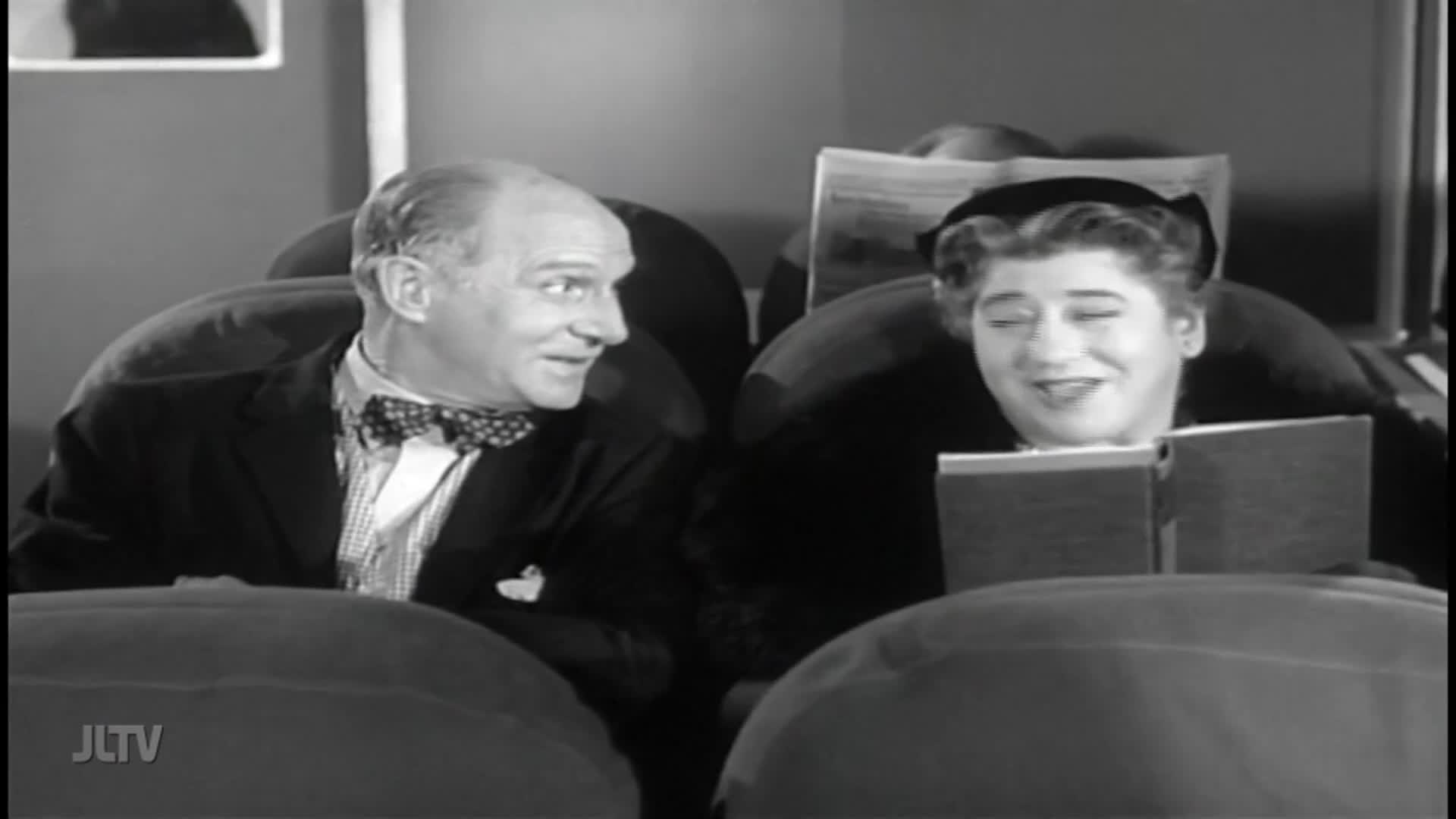A man in a bow tie leans toward a woman, smiling as she reads from a book. The scene, from Jewish Life Television, is set in what looks like a movie theater.
