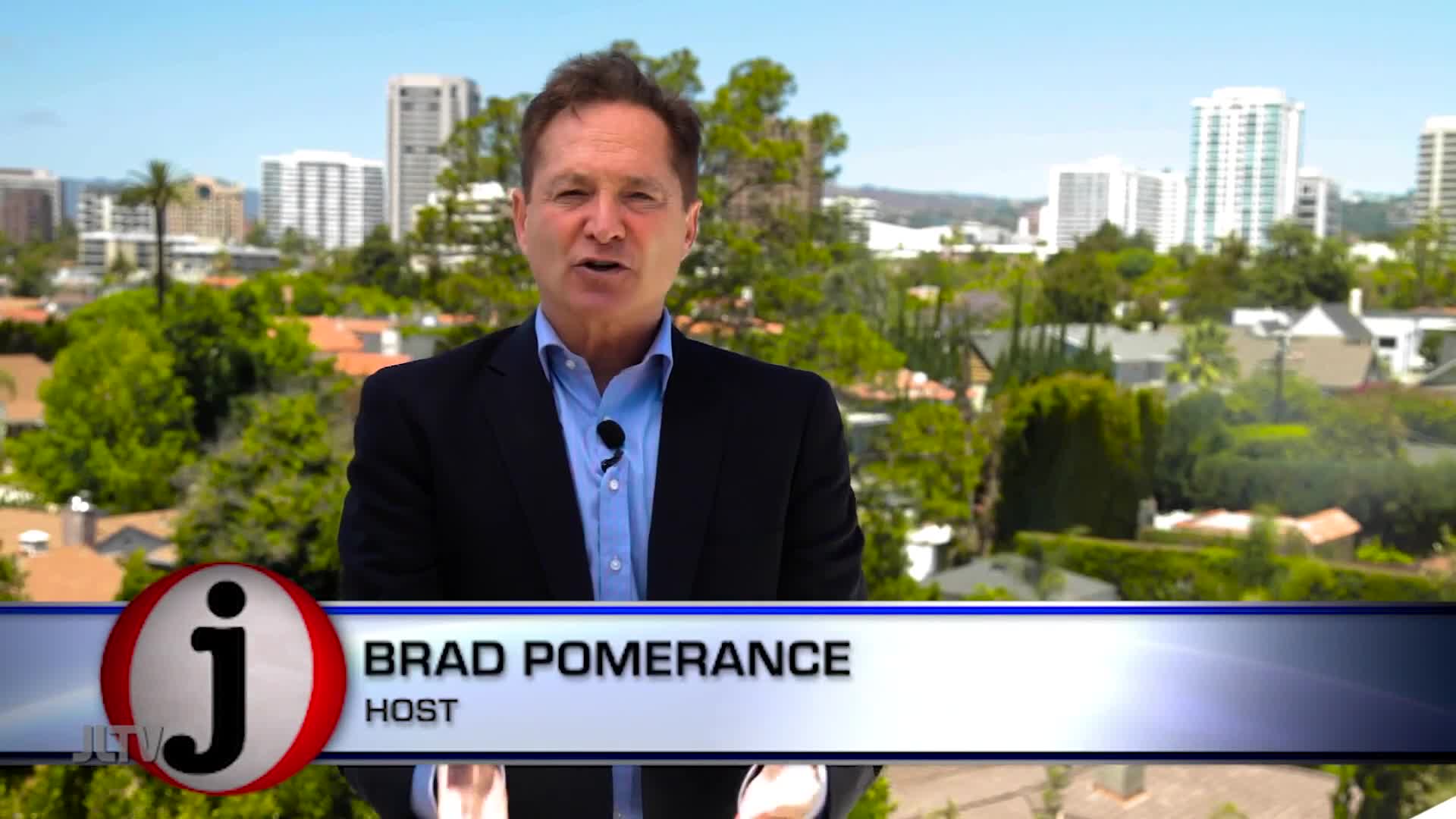 Brad Pomerance, host of a show on Jewish Life Television, is speaking directly to the camera, a microphone clipped to his shirt. Behind him, a sunny cityscape of the United States is visible.
