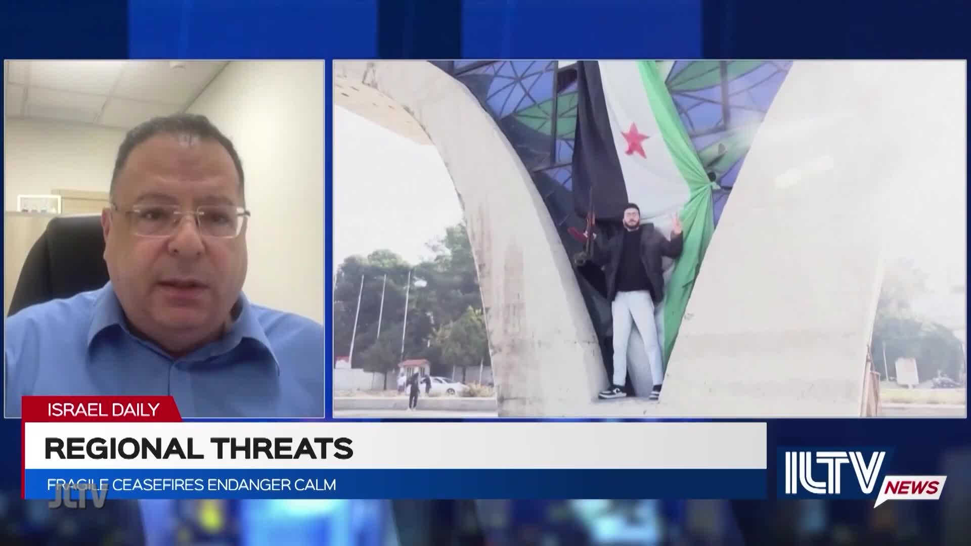 A man in a blazer stands beneath an arch, holding a large flag with a red star. Across the screen, a man in a blue shirt speaks, as the news segment on Jewish Life Television discusses regional threats.
