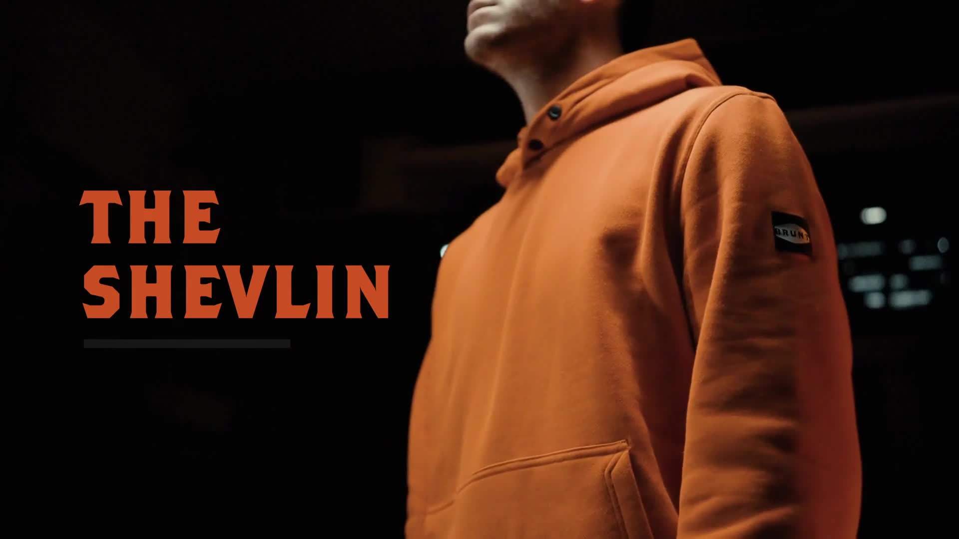 The title "The Shevlin" is displayed in orange, bold lettering against a dark background. A man in an orange hoodie stands in the shadows, his face partially obscured.
