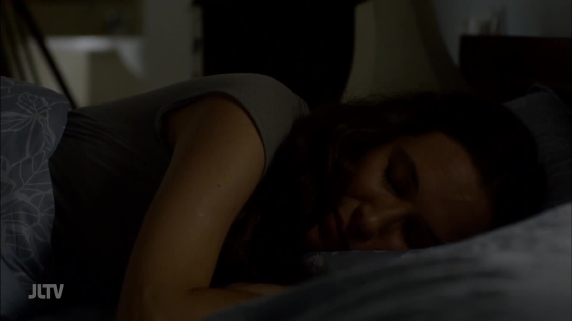 A woman lies asleep, her face pressed into a pillow on a bed. The scene is dimly lit, with the JTLV logo in the corner.
