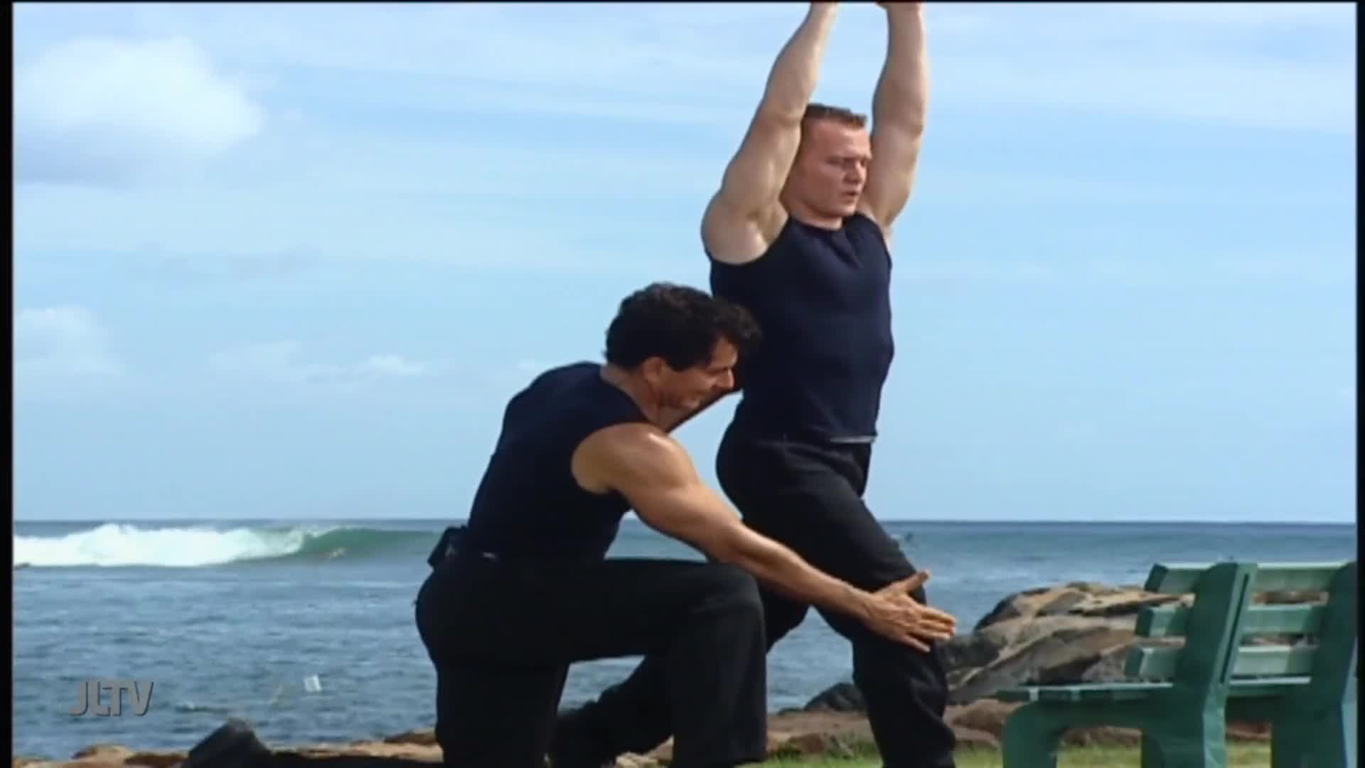 On the beach, a man in a black tank top kneels, guiding another man's leg as he stretches. The second man stands with arms raised, his body angled towards the ocean.
