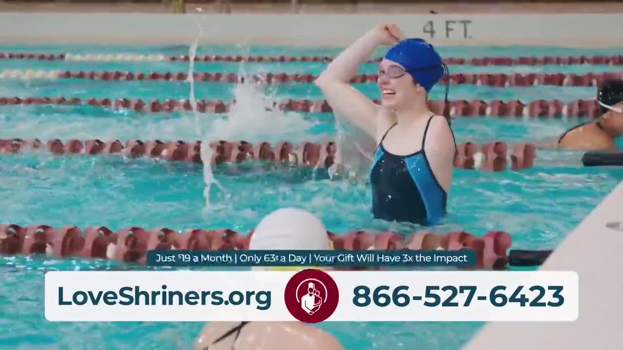 A swimmer in a blue cap raises an arm, water splashing around her as she beams. Other swimmers are in the pool, and the scene is likely part of a commercial for LoveShriners.org, featured on United States Jewish Life Television.
