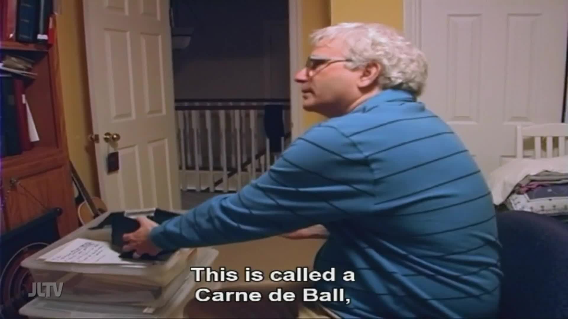 A man in a blue striped shirt sits at a desk, looking to the side. He's holding something black over a box of papers, as the Jewish Life Television logo appears in the corner.
