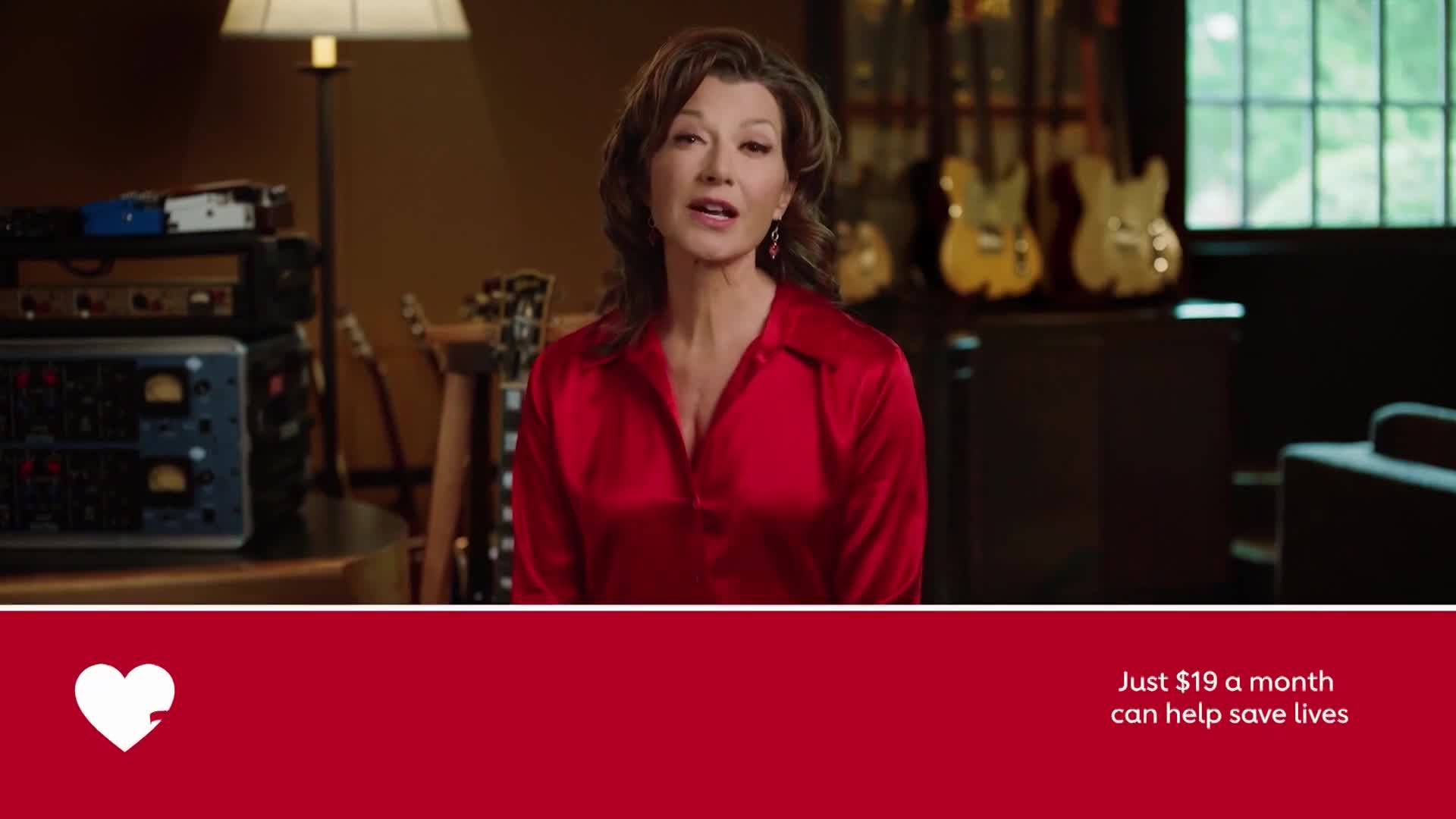 A woman in a red blouse speaks directly to the camera, likely on United States Jewish Life Television. Behind her, guitars and audio equipment are visible in a warmly lit room.
