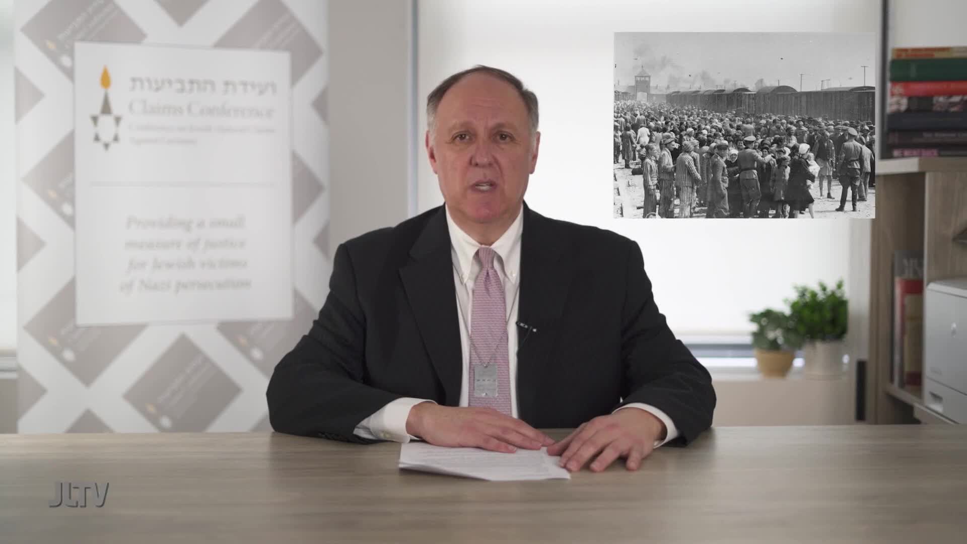 A man in a suit sits at a desk, speaking to the camera for Jewish Life Television. Behind him, a black-and-white image of a crowd of people is displayed.
