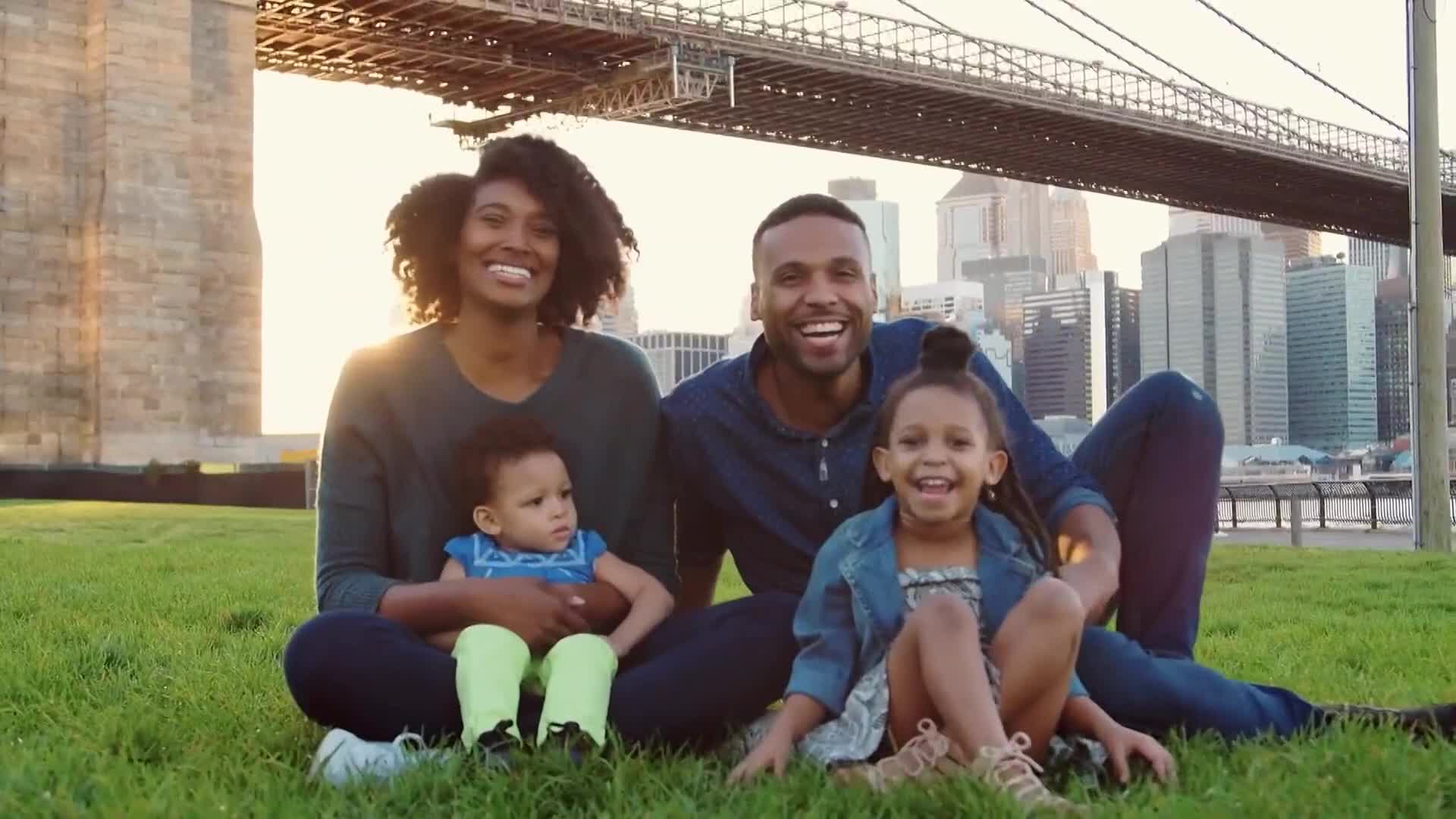 A smiling family sits on the grass, the Brooklyn Bridge looming in the background. The sun shines on their faces as they pose for the camera, looking happy and relaxed.

