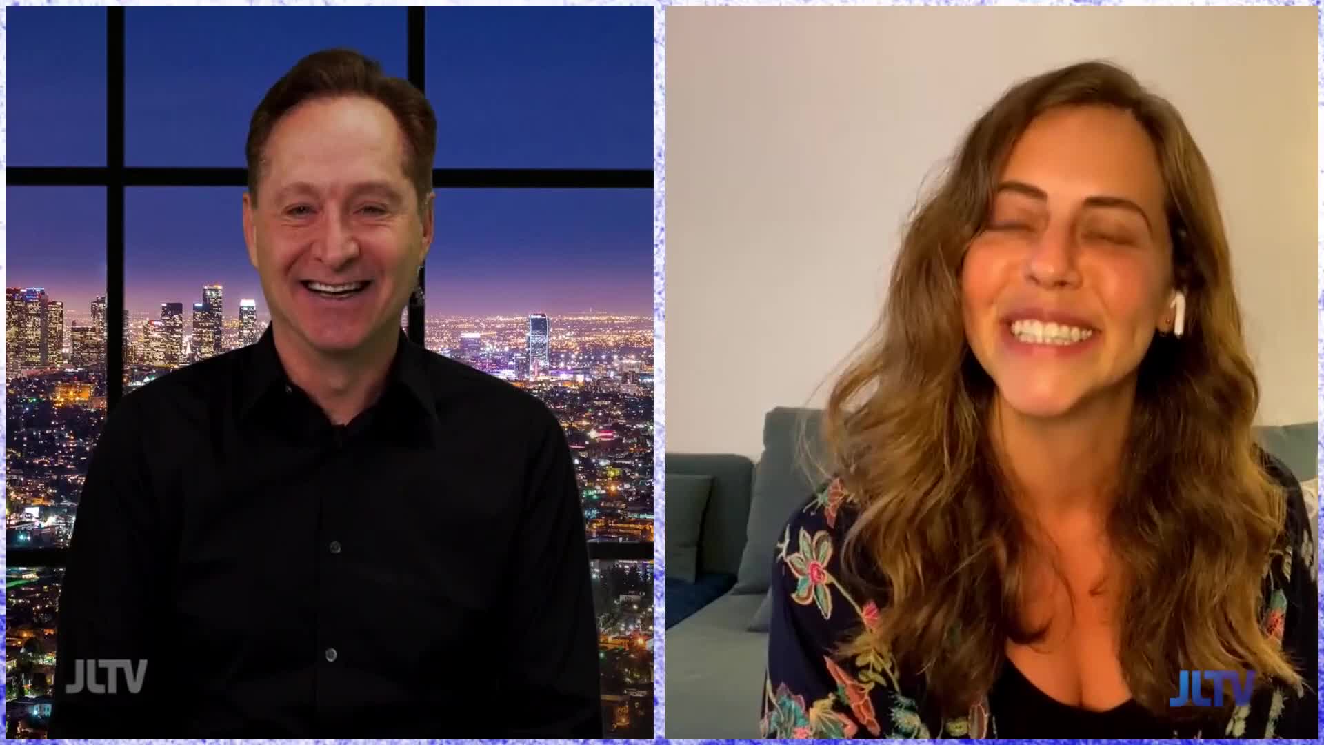 On Jewish Life Television, a man in a black shirt grins, framed against a city skyline. Across the screen, a woman with long hair smiles widely, wearing a floral top and an earbud.
