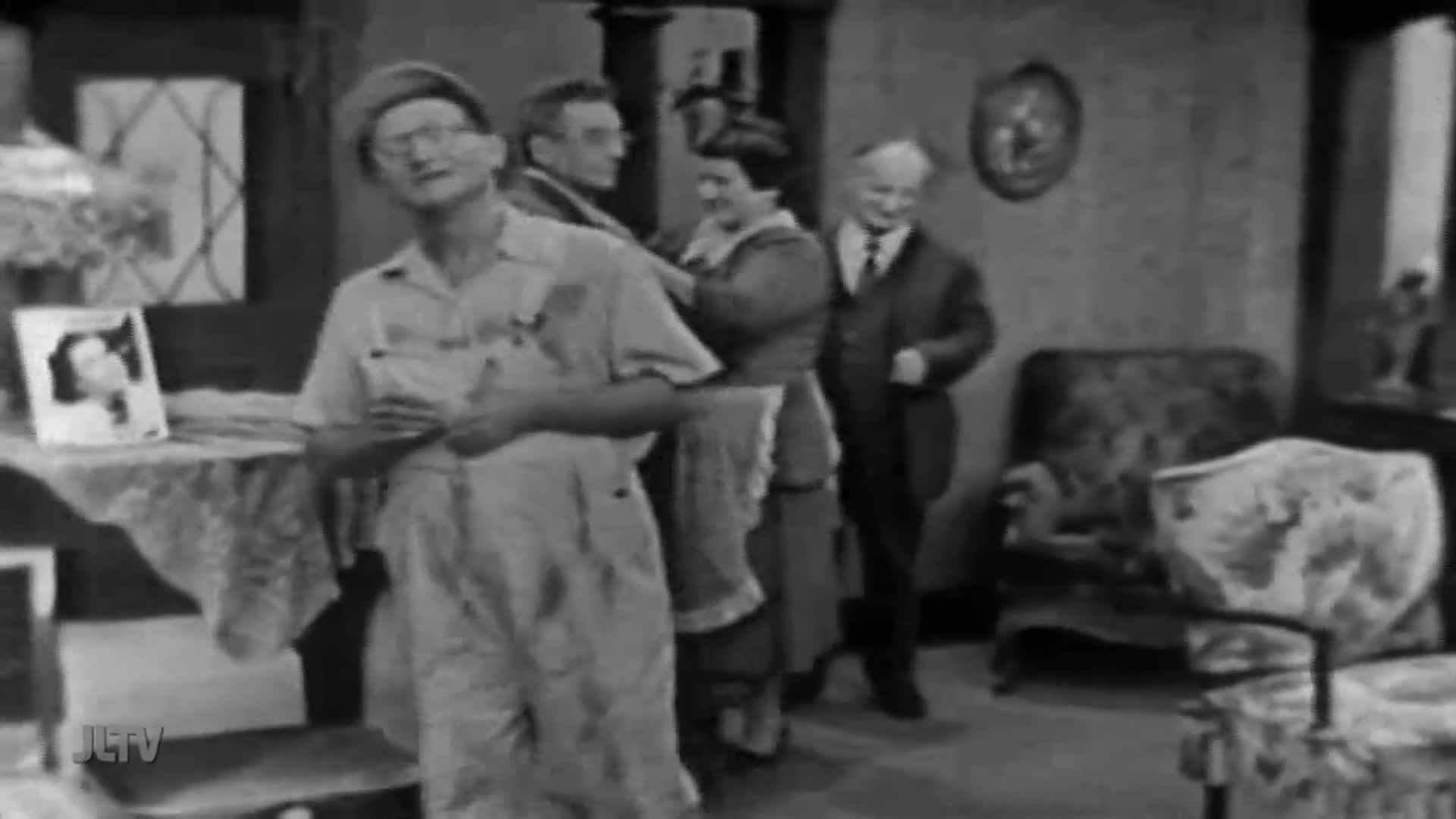 A man in overalls adjusts his suspenders, looking off to the side. Behind him, a woman straightens another man's jacket as a suited gentleman smiles nearby.
