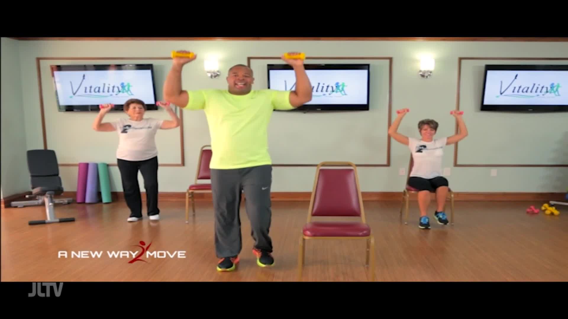 On Jewish Life Television, a man in a bright shirt leads a fitness class, lifting weights with two women. One woman stands, the other sits, both mirroring his movements.
