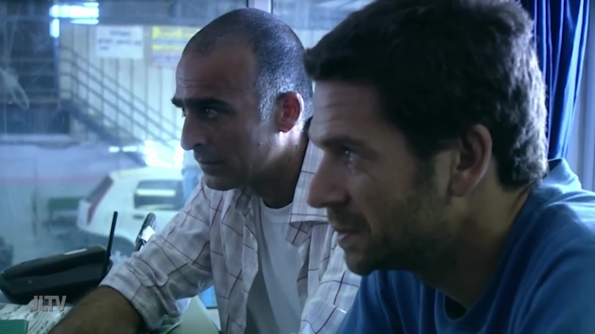 Two men, one in a checked shirt, the other in blue, are focused on something just off-screen. A "JLTV" logo is visible on a desk, and a car is parked outside a window.

