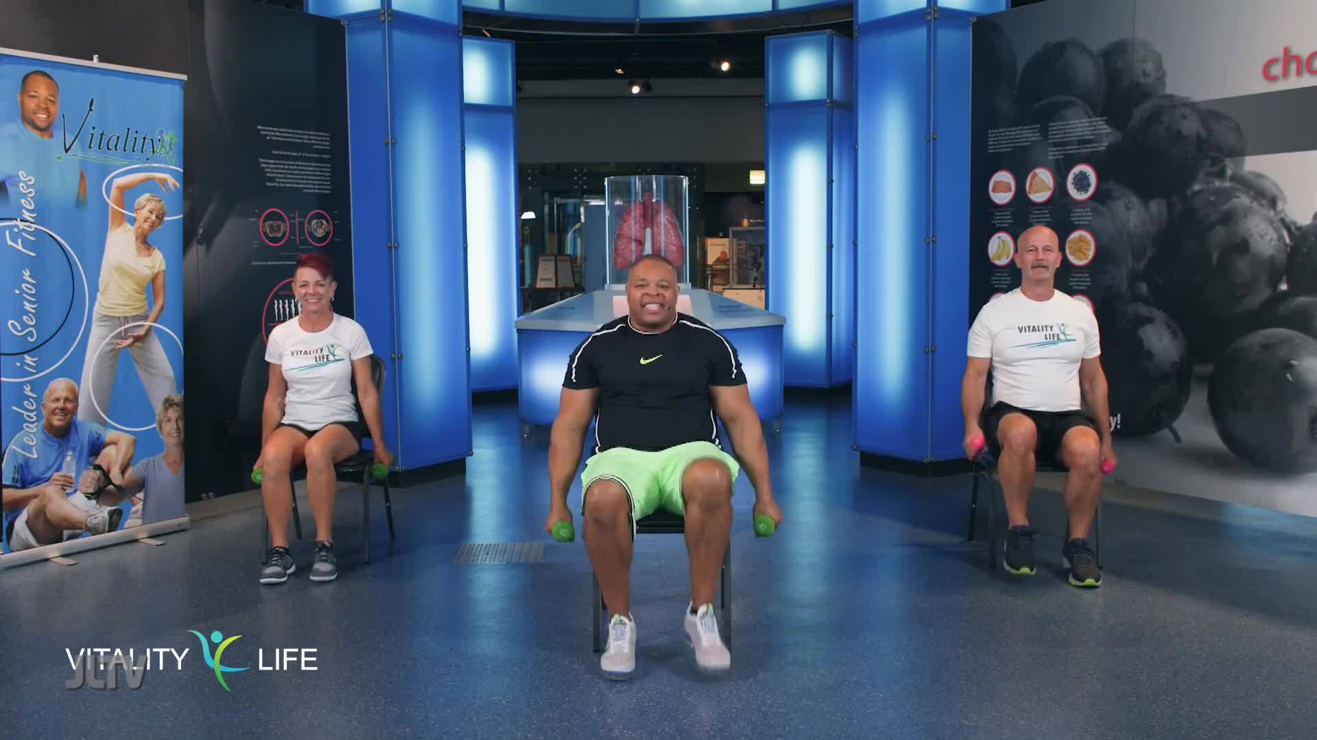 On the set of Jewish Life Television's "Vitality Life," three people are seated, each holding small weights. They're lifting the weights in unison, led by the man in the center.

