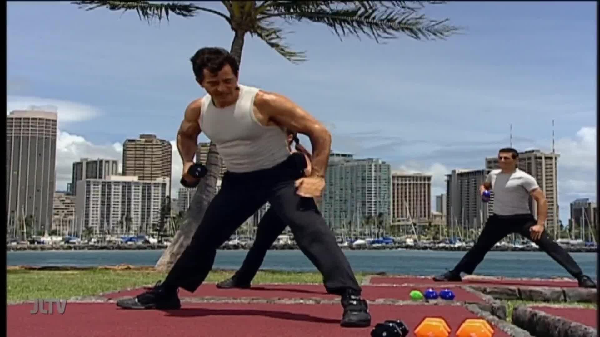 Two men are doing exercises outdoors, with weights in their hands. The camera angle frames the scene against a backdrop of a city skyline near the water, likely a segment on Jewish Life Television.
