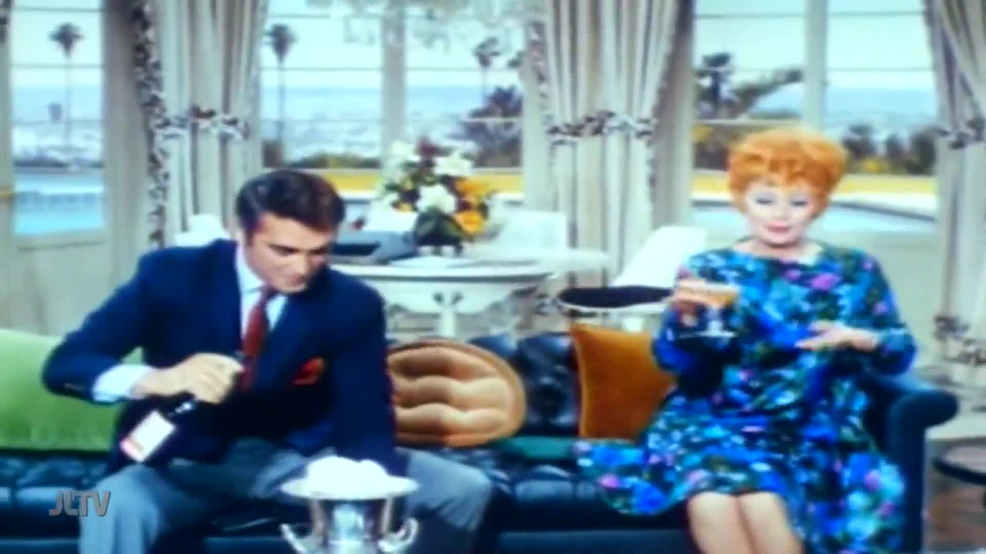 A man in a navy blazer is opening a bottle of wine, while a woman in a floral dress holds a cocktail. They're sitting on a leather couch, looking relaxed, on what appears to be a United States television program, possibly on Jewish Life Television.
