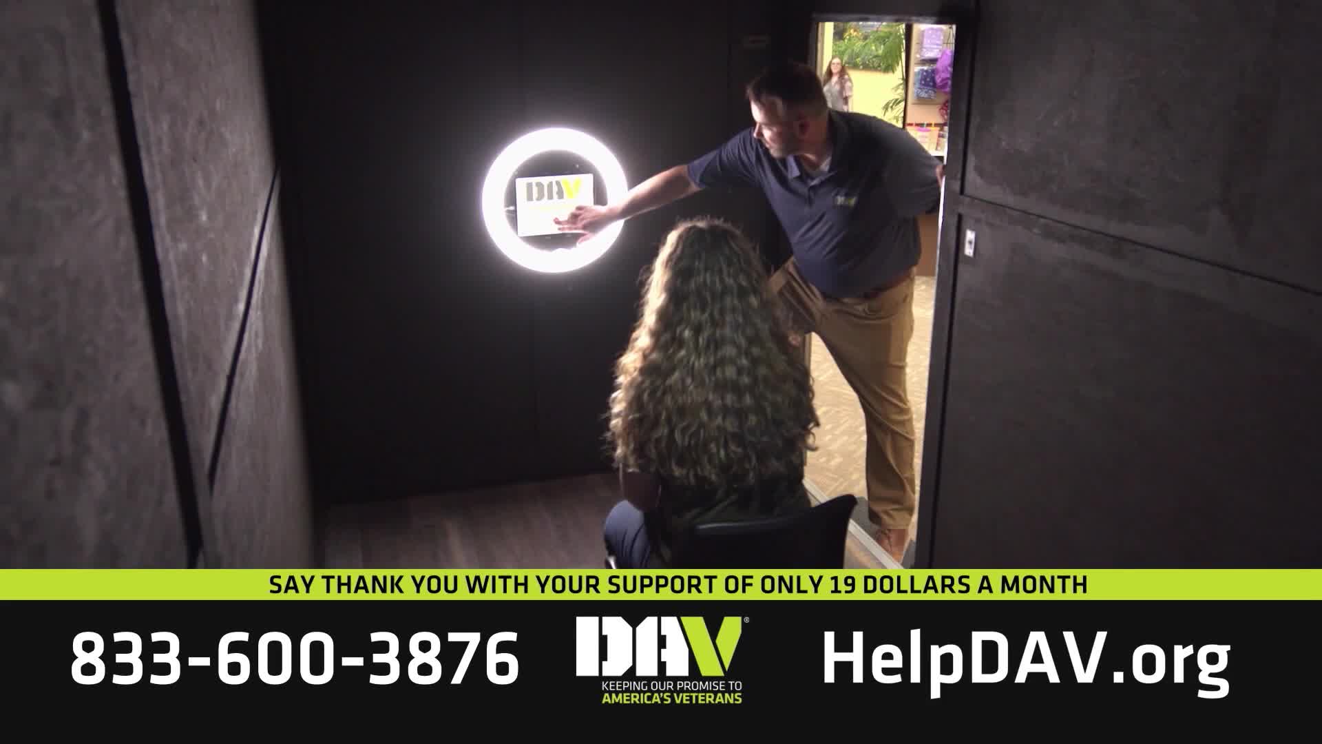 A man in a blue polo shirt and khaki pants leans in to adjust a sign held by a woman with long, curly hair. The ring light behind them illuminates a small white card with the DAV logo.