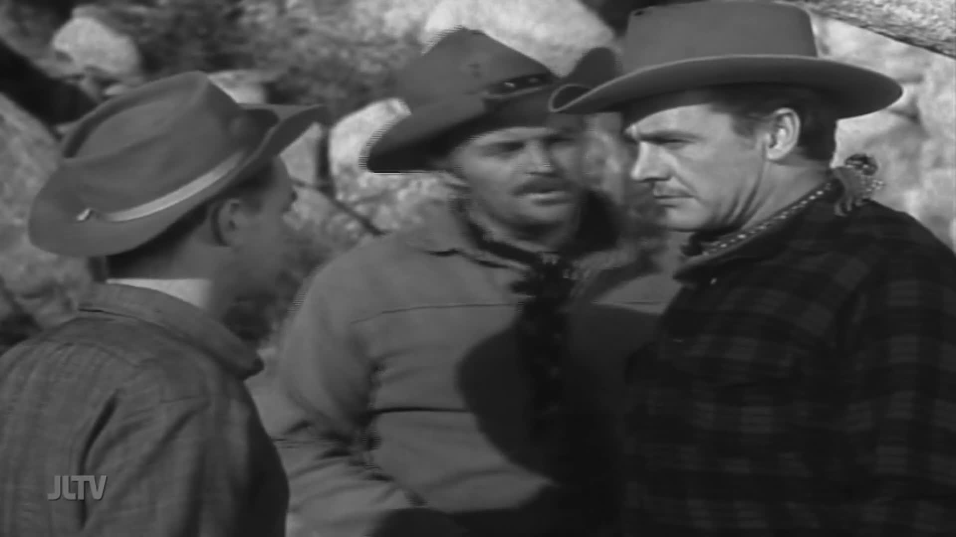 Three men in cowboy hats stand close, their faces turned toward each other. A small monkey perches on the shoulder of the man in the plaid shirt.