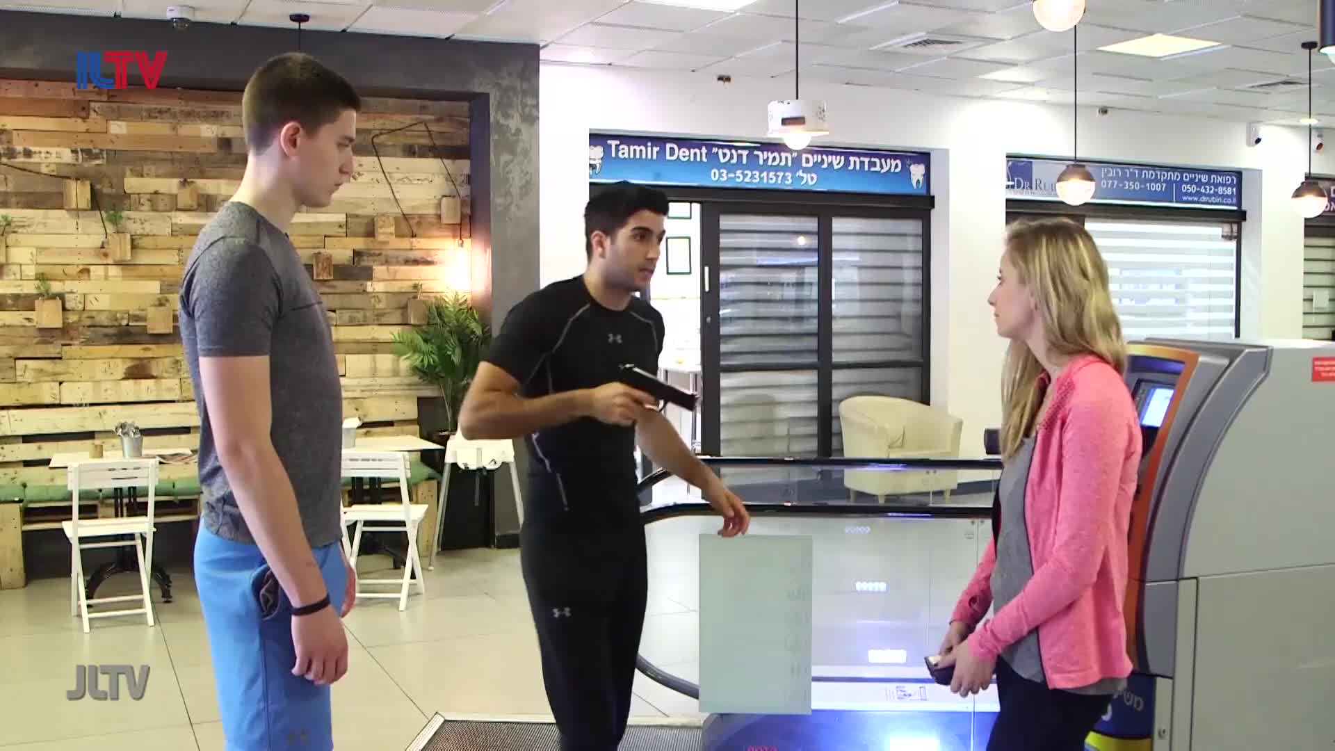 A man in a black shirt points a handgun at another man in a grey shirt. A woman in a pink cardigan stands near an ATM, looking away.