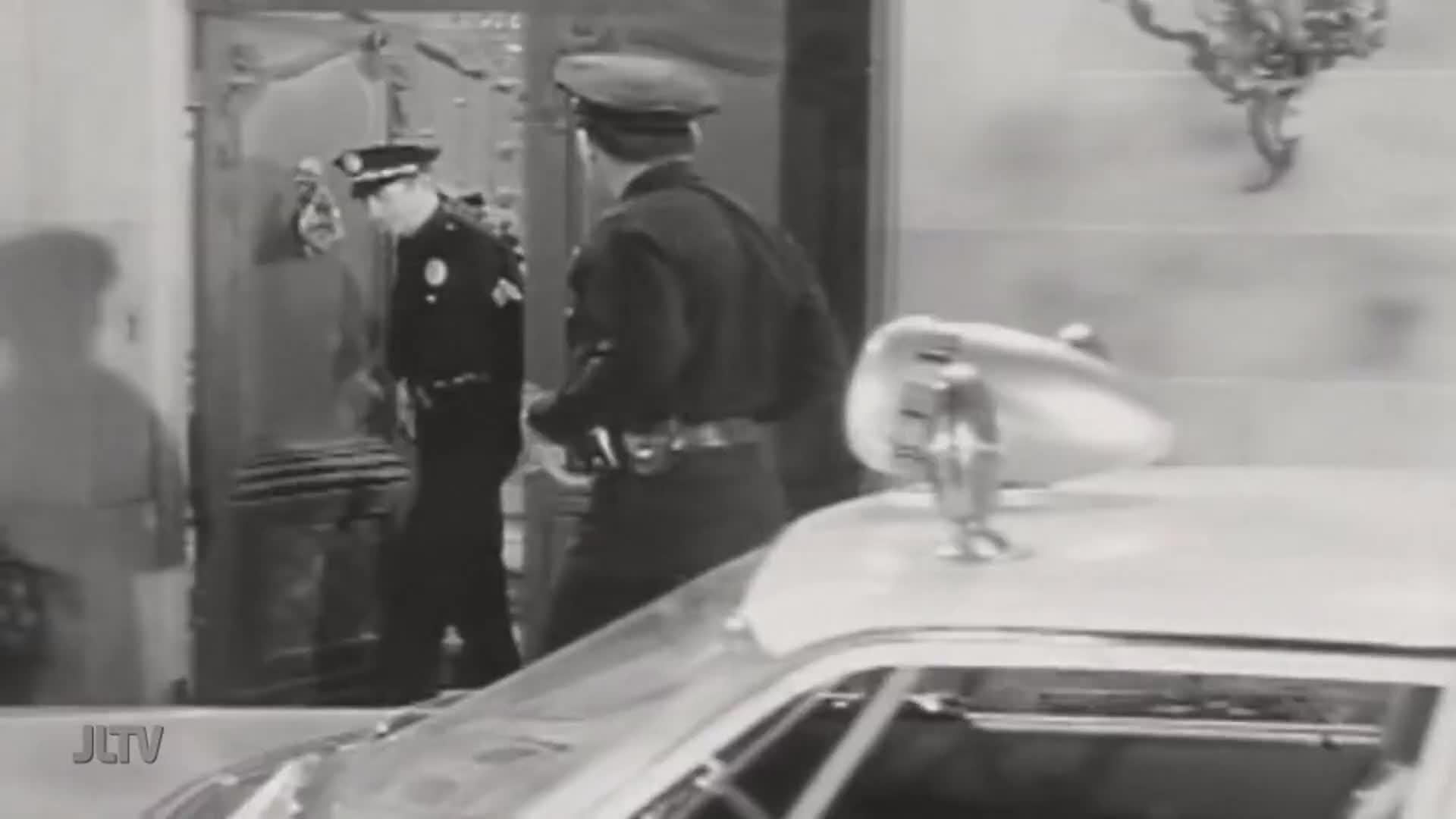 Two police officers in uniform approach a large, ornate doorway. A police car with its siren light is parked in the foreground.