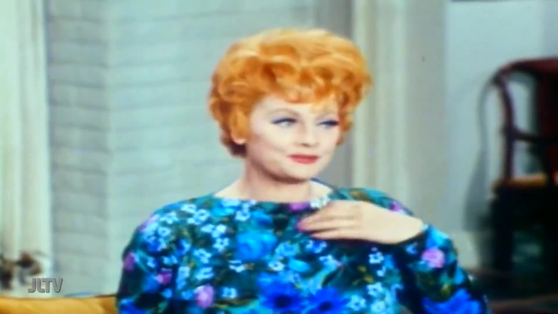 A woman with bright red, voluminous hair sits in a blue floral dress. Her hand rests near her chest as she looks off to the side.