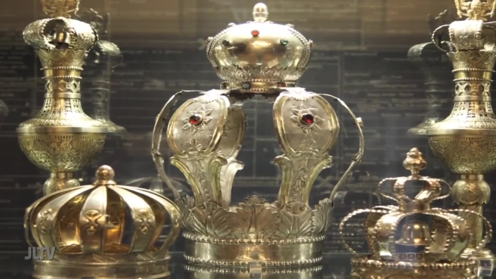 Several ornate crowns gleam under the lights, each a testament to intricate craftsmanship. One prominent silver crown, adorned with red jewels, sits centrally, flanked by golden vessels and smaller crowns.