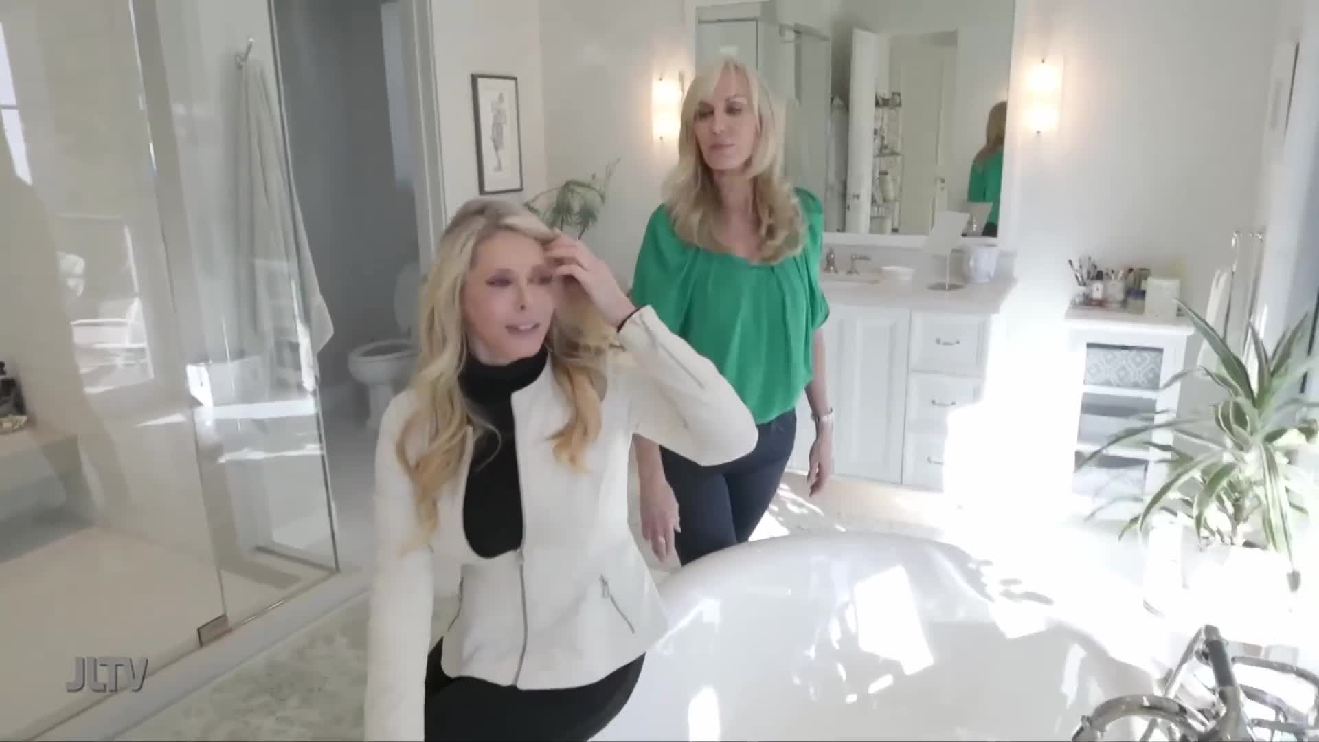 Two women stand in a bright, modern bathroom. One woman, wearing a white jacket, gestures with her hand as she speaks. The other, in a green top, stands behind her, looking towards the camera.