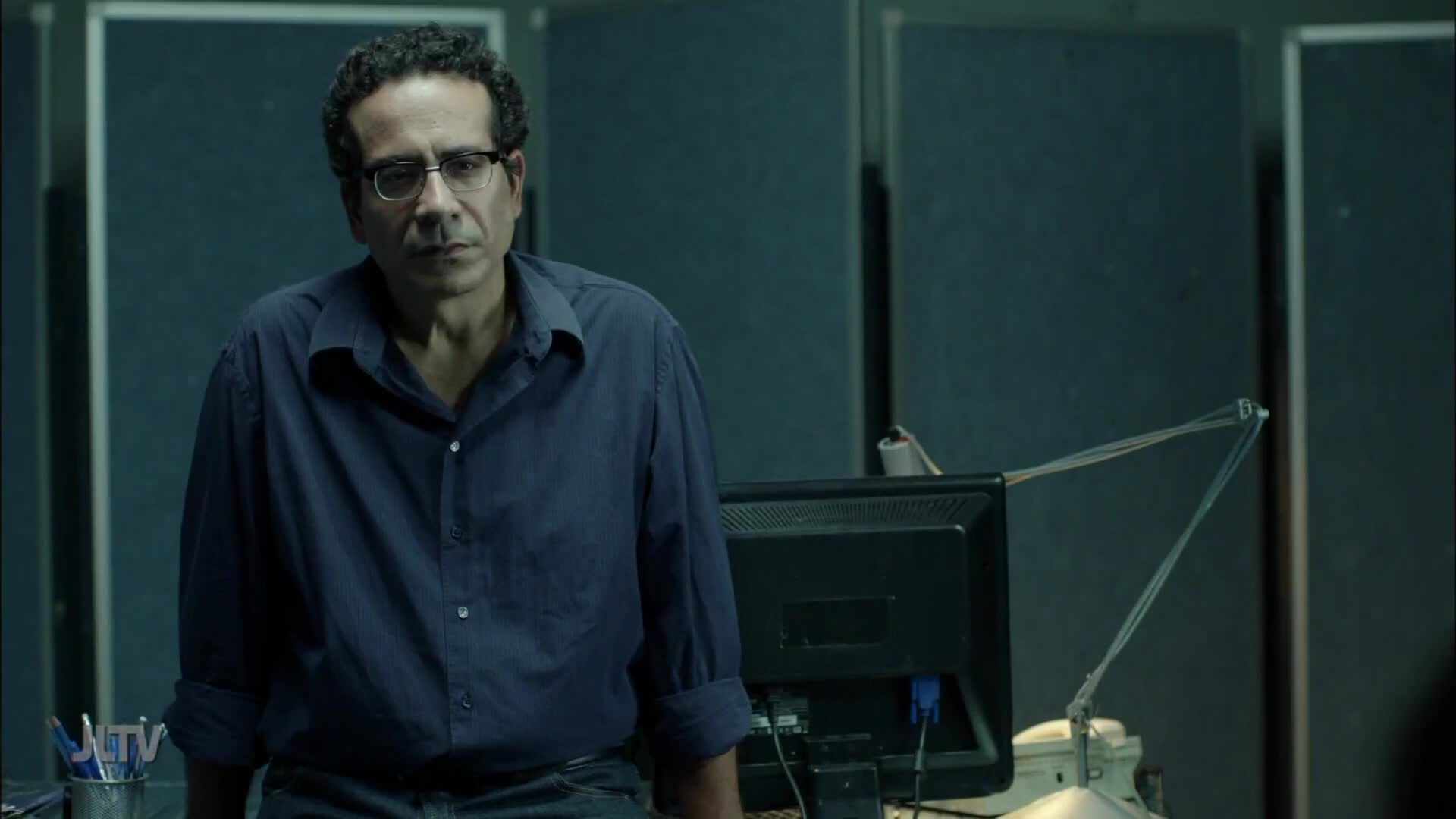 A man with glasses and a blue shirt sits on a desk, looking directly ahead. A computer monitor and a metal cup with pens rest on the surface in front of him.