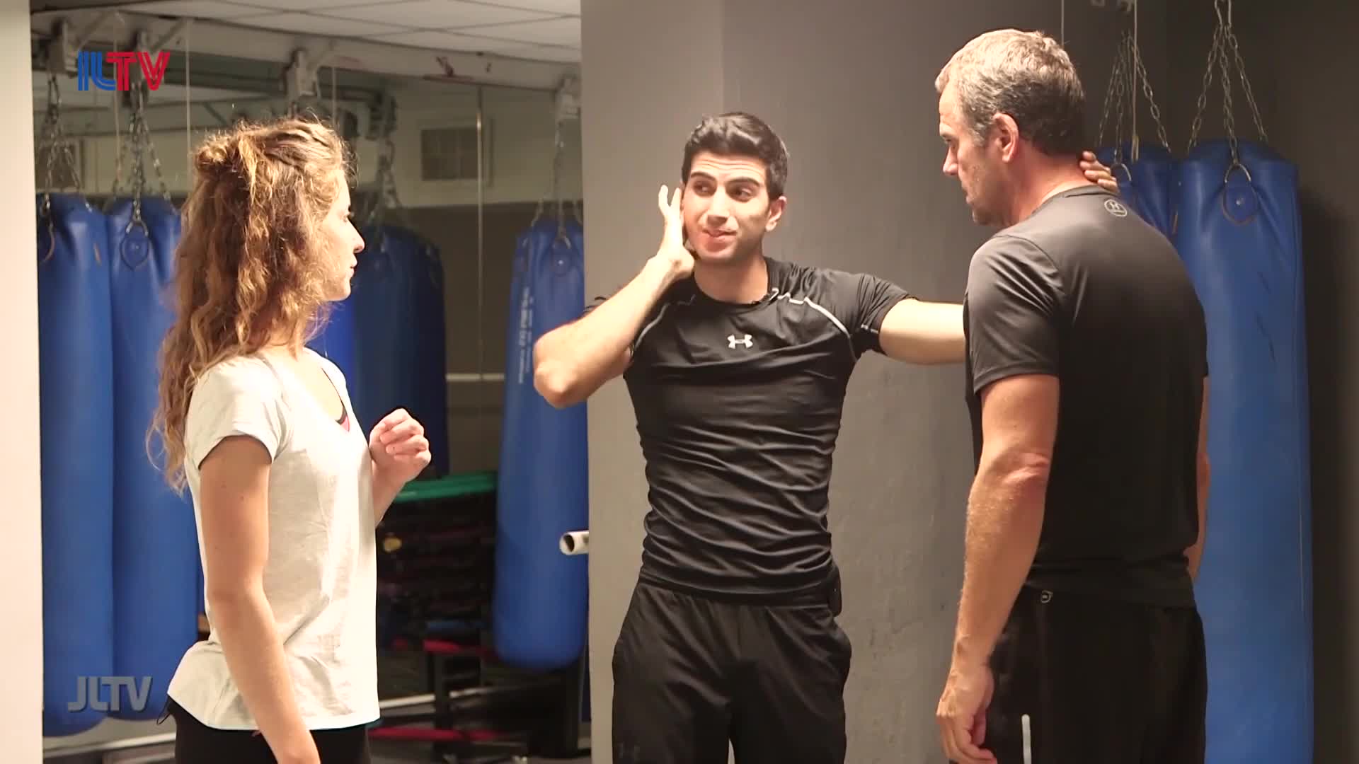 A young man in a black shirt demonstrates a technique, his hand near his ear. A woman in a white t-shirt watches intently, and a man in a dark shirt stands to the side.