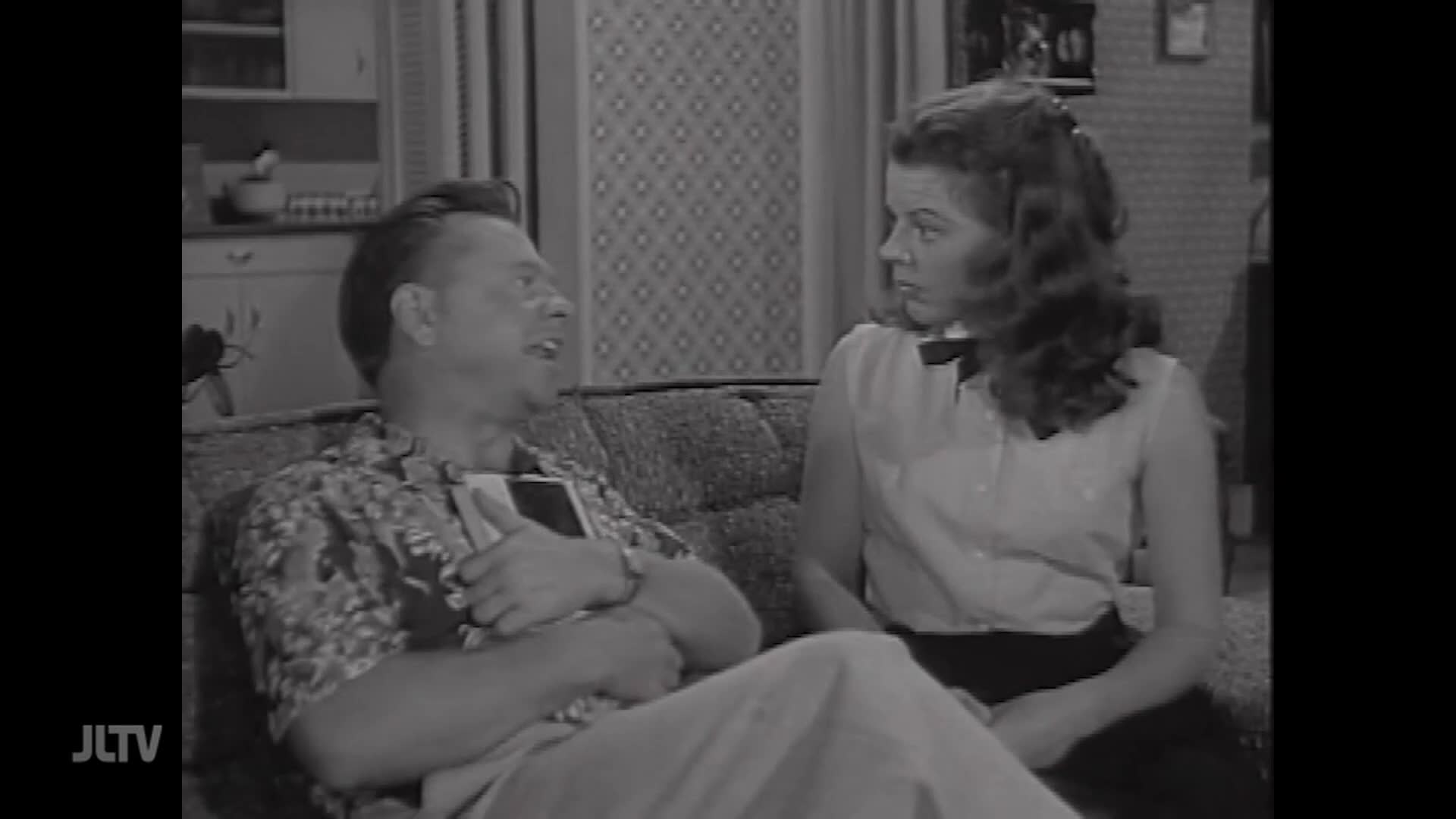 A man in a patterned shirt gestures with a book in his lap while talking to a young woman in a collared shirt. They sit on a couch in what appears to be a living room.