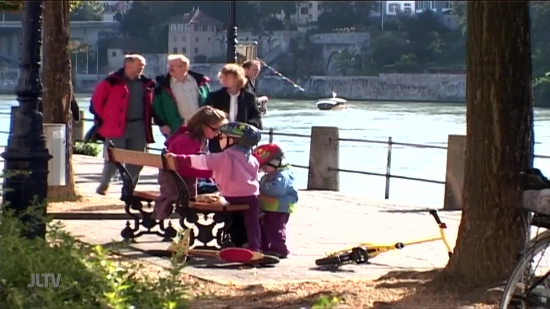 A group of children huddle near a park bench, their helmets bright against the dappled sunlight. A boat glides across the river in the background.