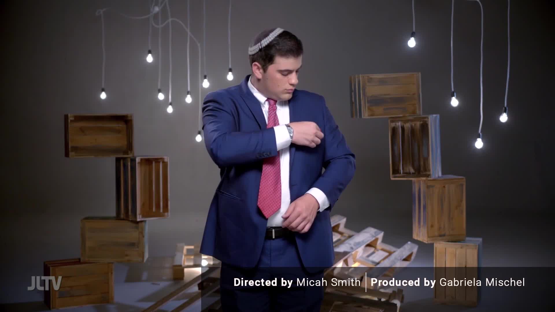 A young man in a suit and kippah adjusts his jacket. He stands amidst a collection of wooden crates and bare lightbulbs hanging from wires.
