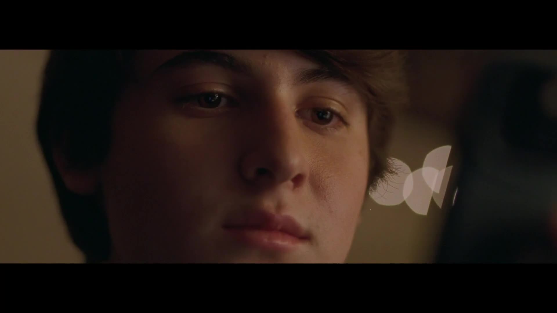 A young person looks down, eyes focused on something out of frame. Soft, circular lights drift in the background, hinting at a warm, indoor setting.