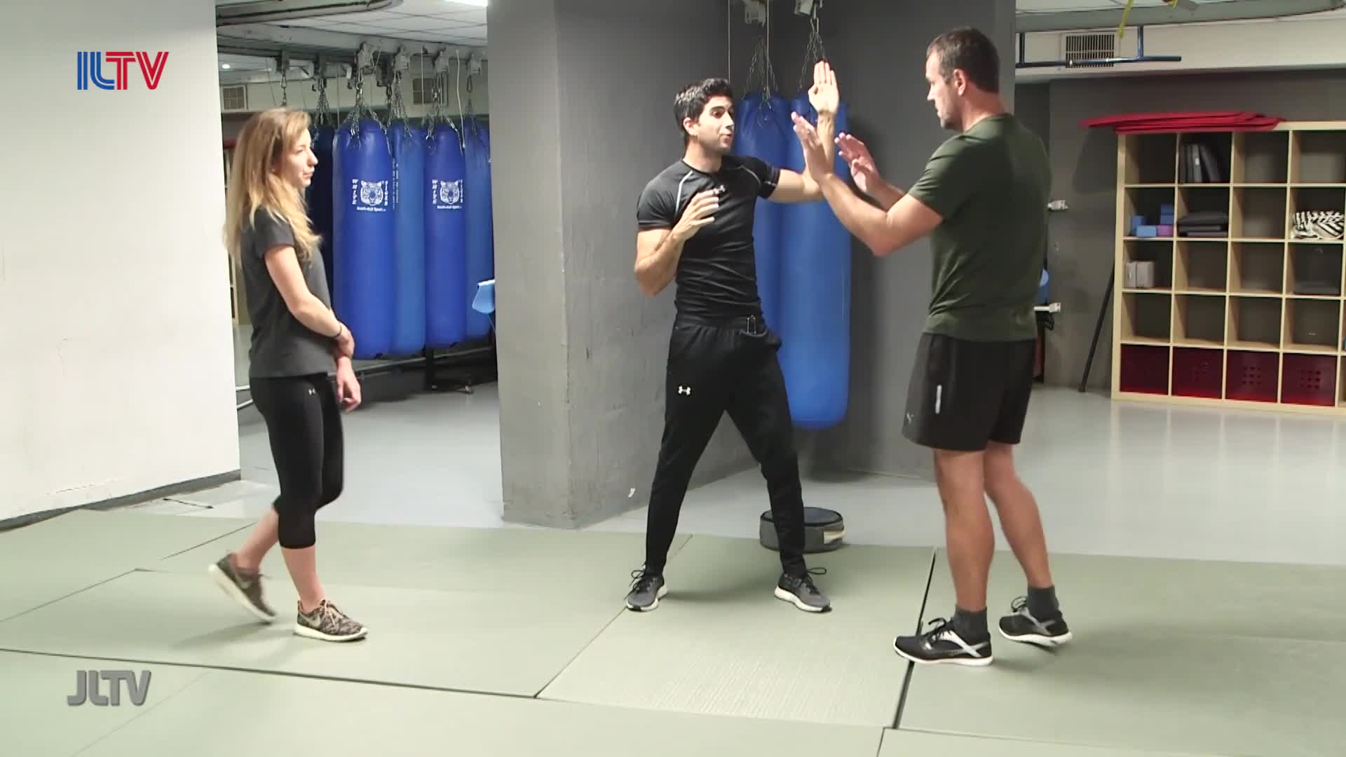 A man in a black shirt and pants demonstrates a defensive move, hands raised, while another man in a green shirt mirrors his stance. A woman in athletic wear watches them from the side.