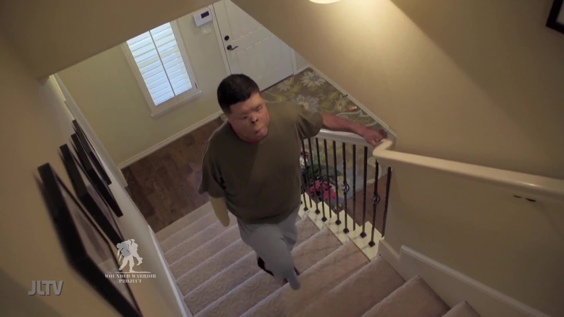 A man with a prosthetic arm is climbing a carpeted staircase, his hand gripping the banister for support. The scene is part of a segment for Jewish Life Television, highlighting the work of The Yeshiva Warrior Project.