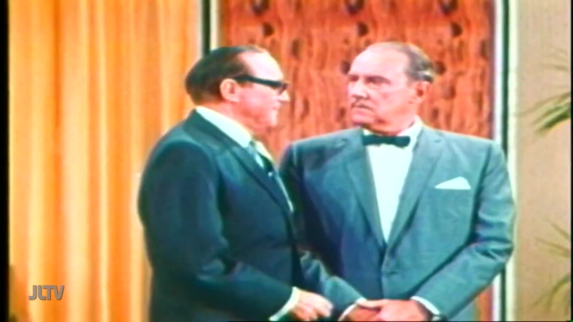 Two men in suits stand facing each other, one with dark glasses, the other with a bow tie. Their hands are clasped in a handshake.