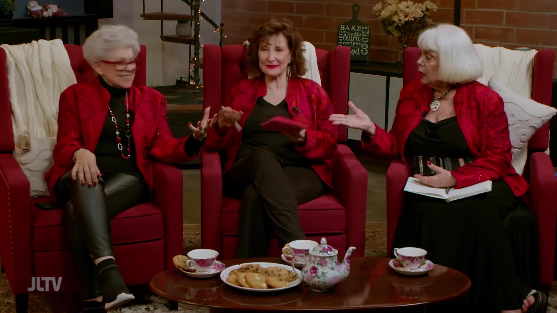 Three women, dressed in festive red jackets, are seated in plush chairs, engaged in conversation. A tea set and cookies sit on a low table between them, suggesting a relaxed gathering.
