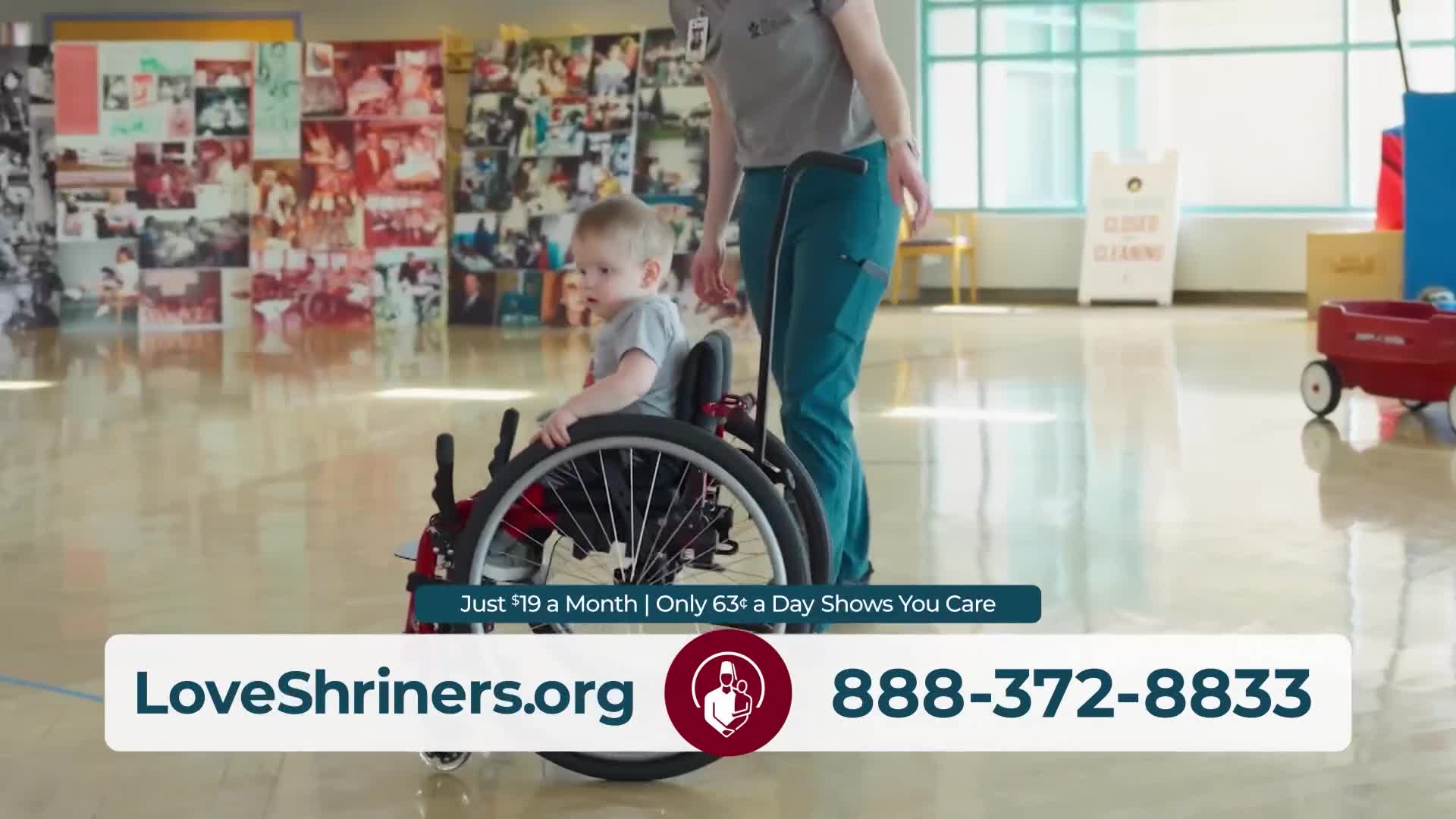 A woman in teal scrubs pushes a young boy in a wheelchair across a polished floor. A wall covered in photos stretches behind them.