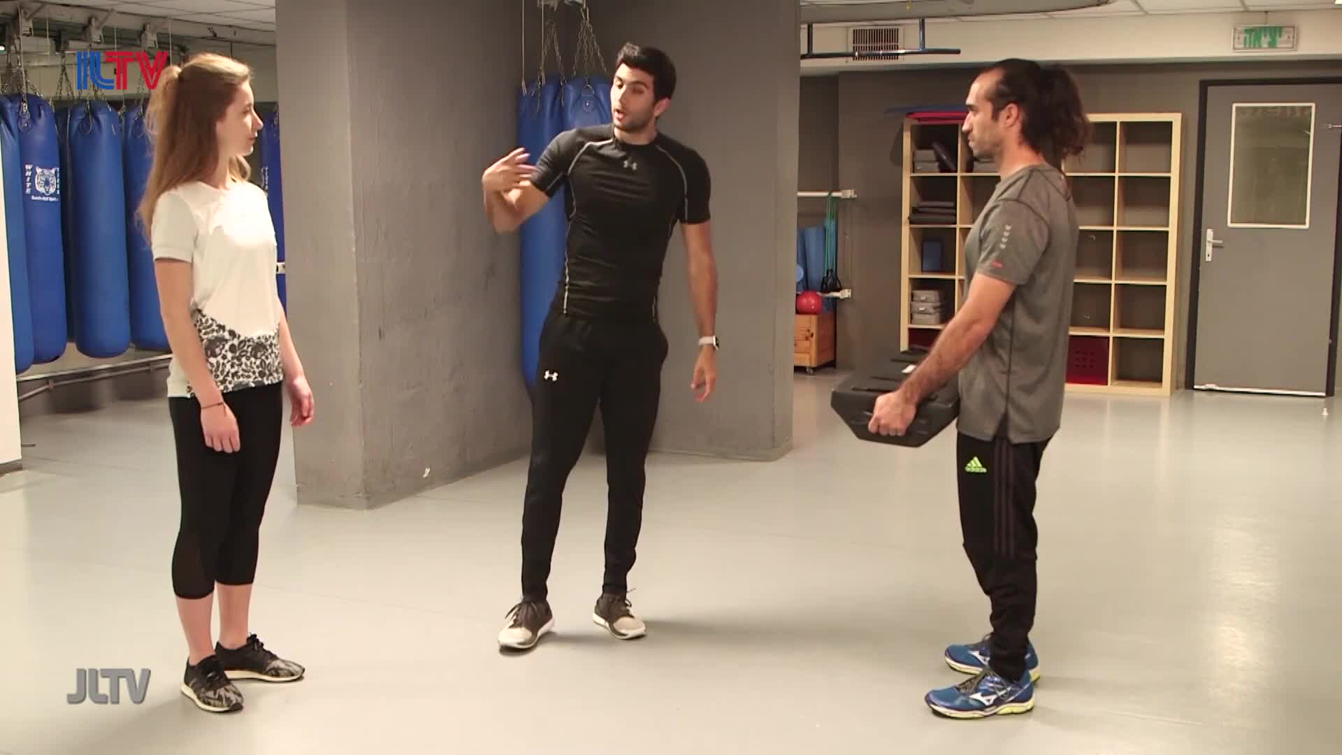 A man in a black shirt demonstrates a technique, gesturing with his hands. Another man stands opposite him, holding a black weight. A young woman watches them, her hands clasped in front of her.