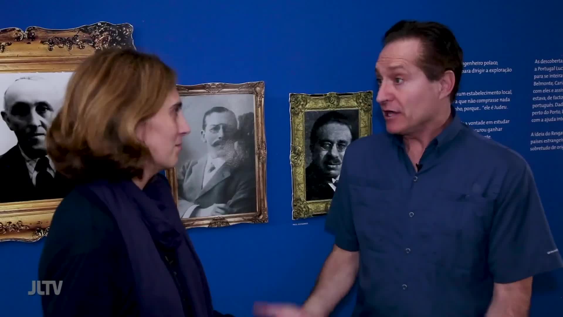 A man in a blue shirt gestures as he speaks, facing a woman in a dark jacket. Behind them, framed portraits hang on a vibrant blue wall, some featuring men with formal attire and mustaches. The JLTV logo appears in the lower left corner.
