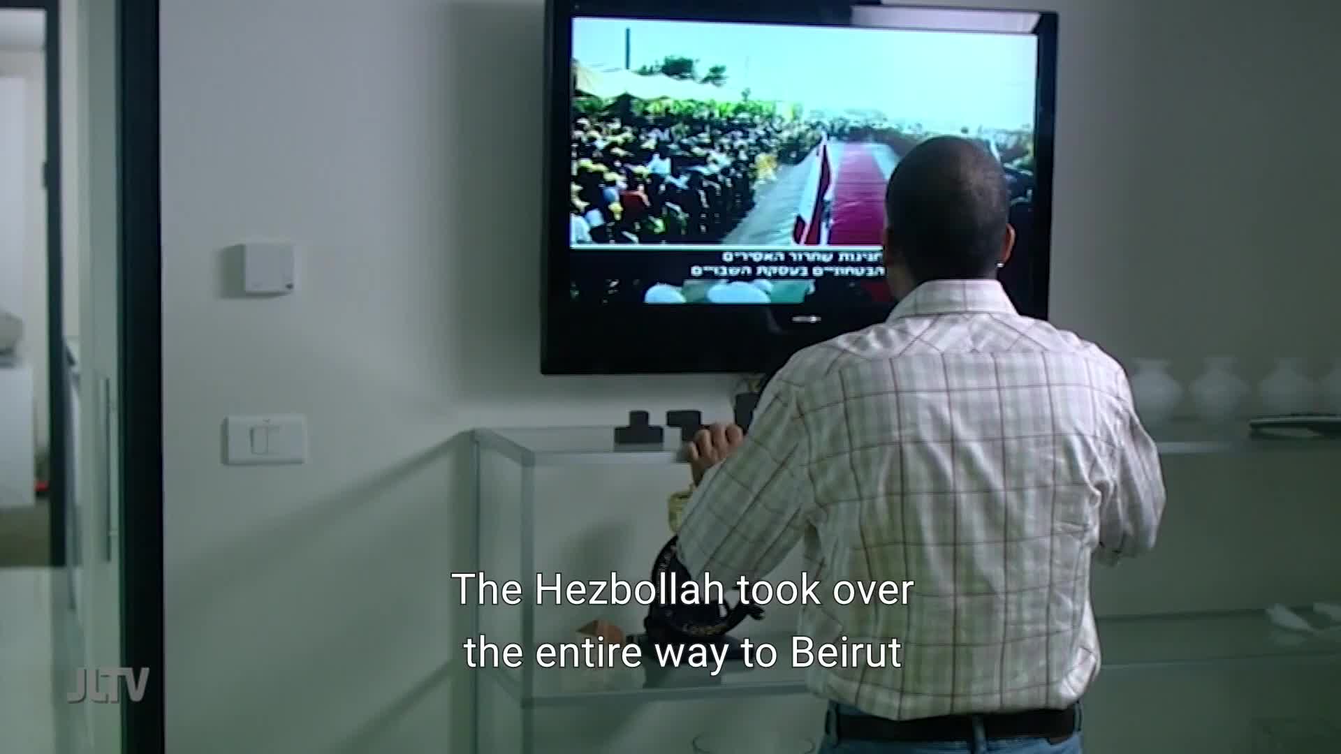 On the television, a large crowd gathers along a red carpet. A man in a plaid shirt stands before the screen, his back to me. The text on the screen reads, "The Hezbollah took over the entire way to Beirut."
