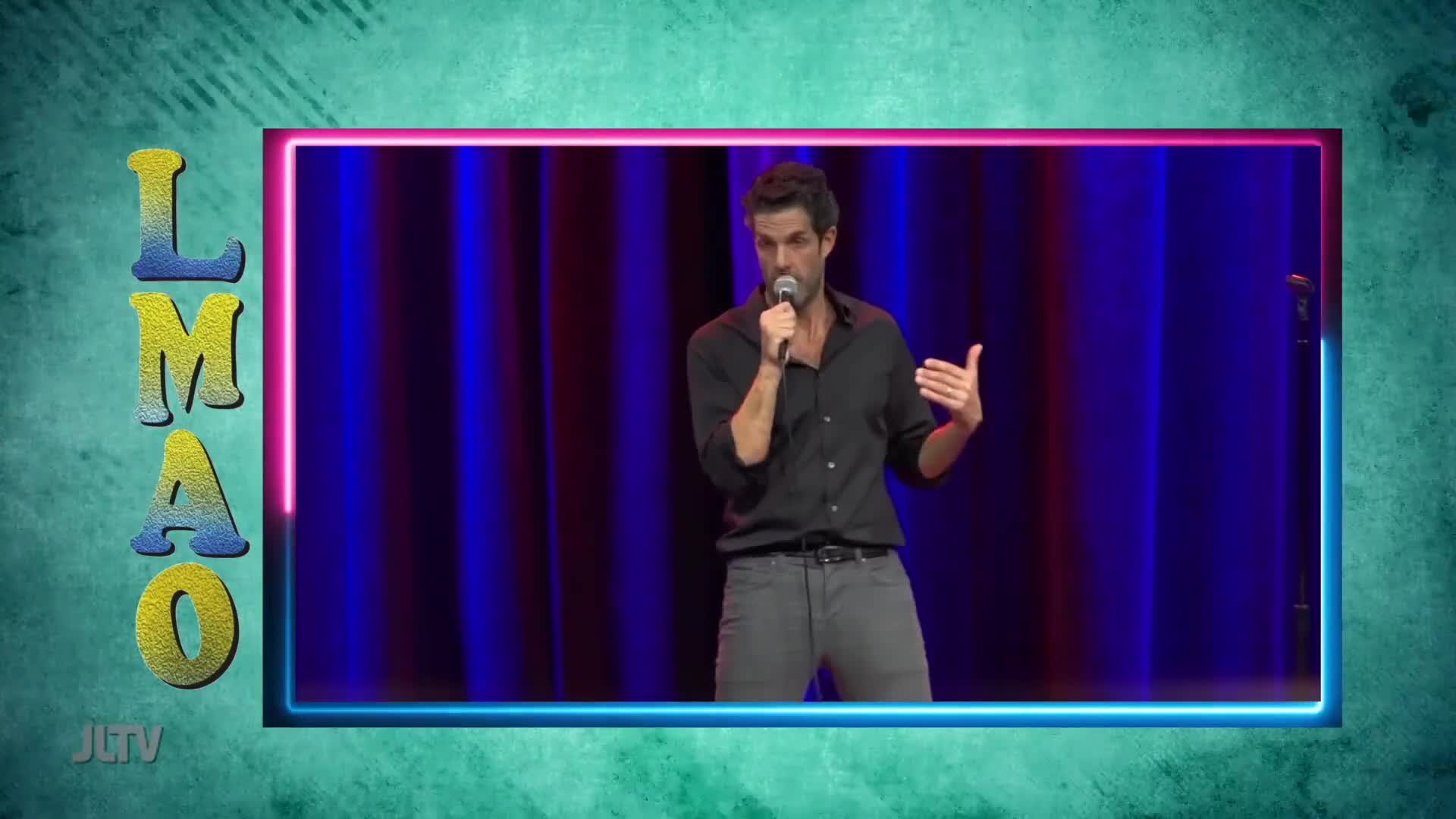 A man holds a microphone and gestures with his left hand while speaking on a stage. The backdrop is a deep blue curtain, framed by neon pink and blue lights.