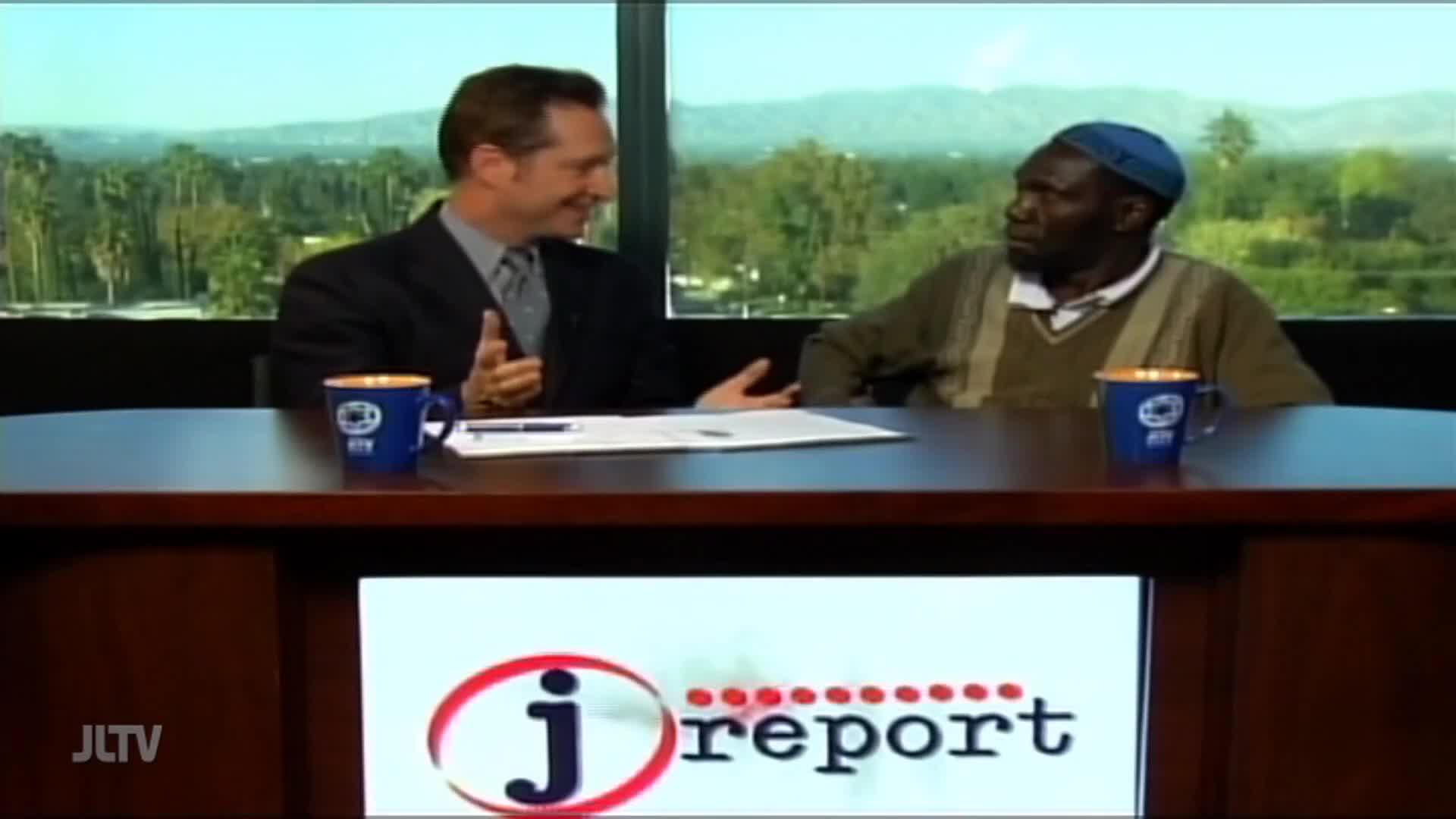 Two men sit at a table, engaged in conversation. The man on the left, wearing a suit, gestures with his hands while the man on the right, in a sweater and cap, listens intently. A blue mug with a logo sits on the table between them, and behind them, a window offers a view of trees and distant hills.
