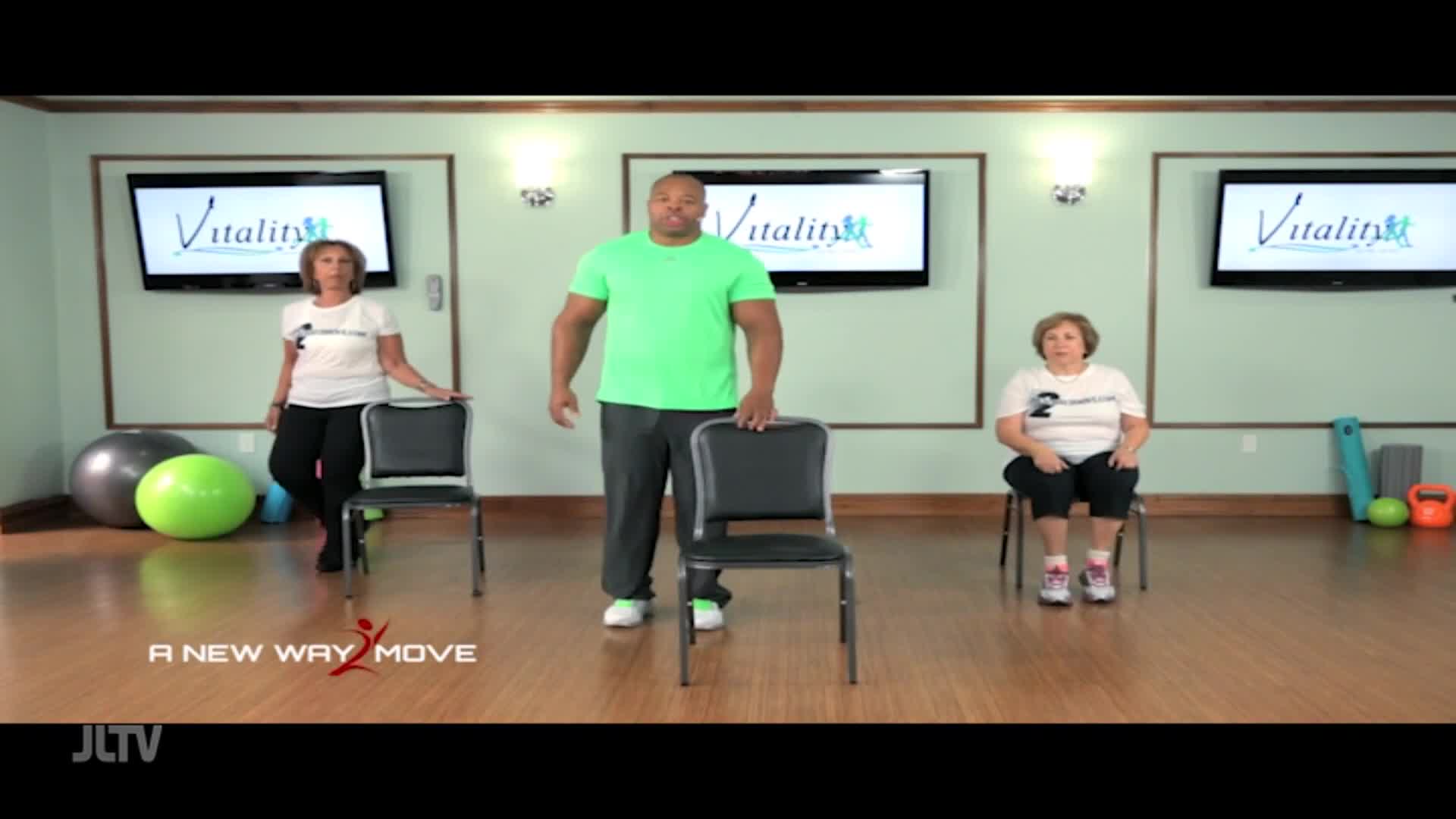 A man in a bright green shirt stands by a chair, while two women, one standing and one seated, are positioned on either side of him. The screen behind them displays "Vitality" alongside a logo.
