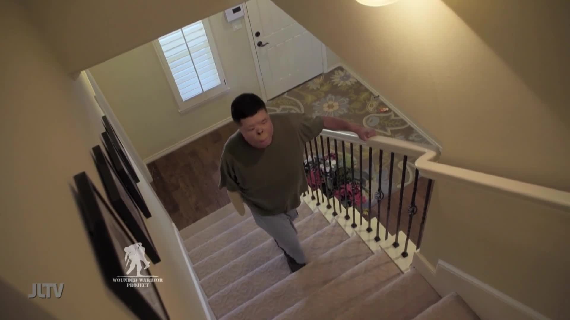 A man ascends the carpeted stairs, his hand resting on the banister. The JLTV logo appears in the corner, alongside the Wounded Warrior Project emblem.