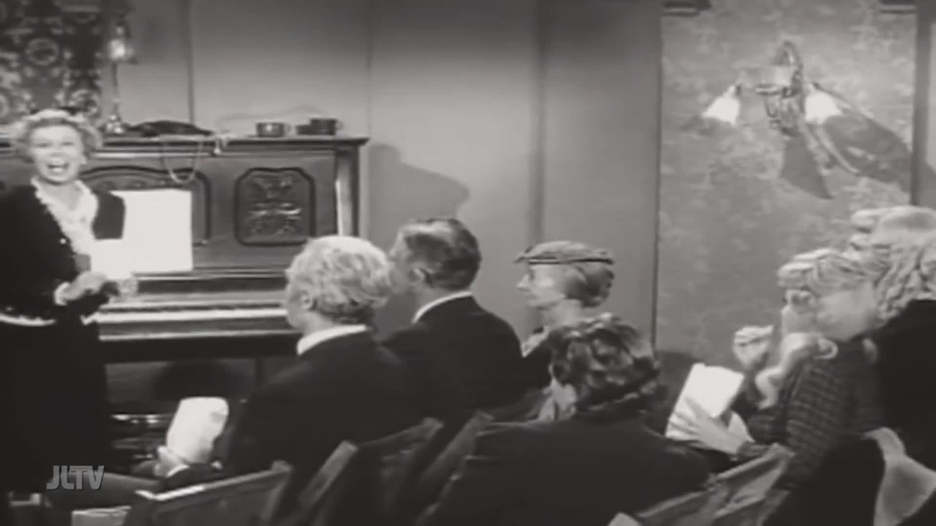 A woman stands by a piano, holding a sheet of music and smiling broadly at the assembled audience. Several people in the foreground are seated, facing her, some holding papers.