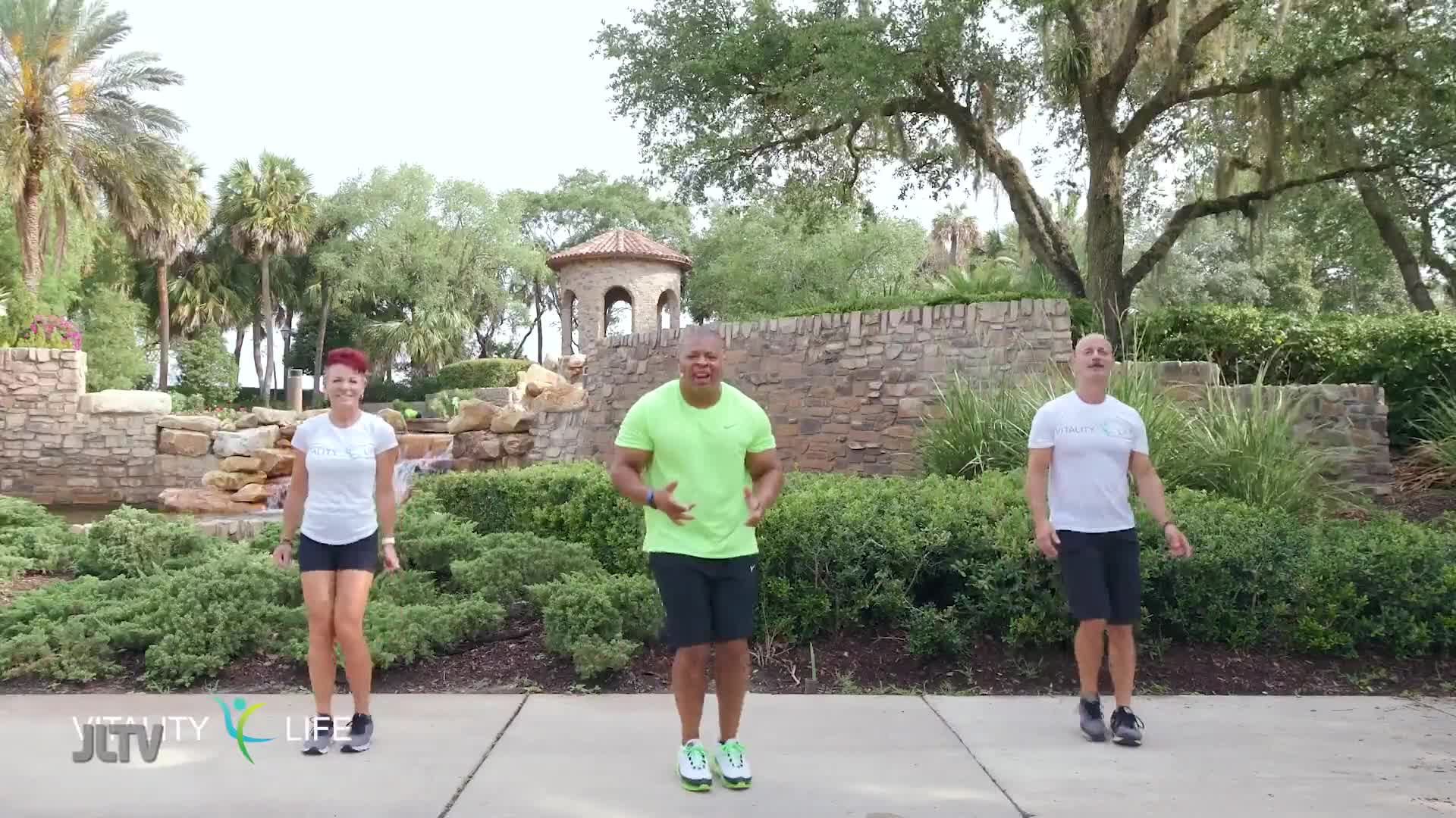 Three people are jumping in place on a paved path. A woman with short red hair and a white shirt is on the left, a man in a bright green shirt is in the middle, and a man in a white shirt is on the right.