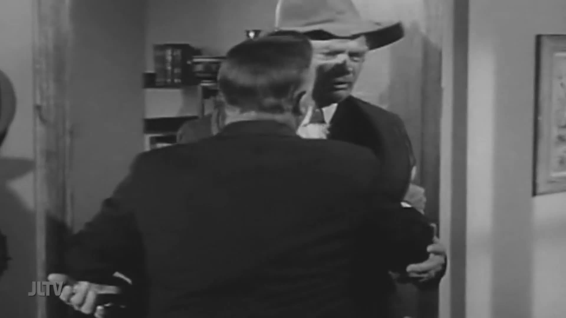 A man in a suit and fedora stands facing another man in a dark suit. The man in the fedora is speaking, his mouth open, as the other man turns away.