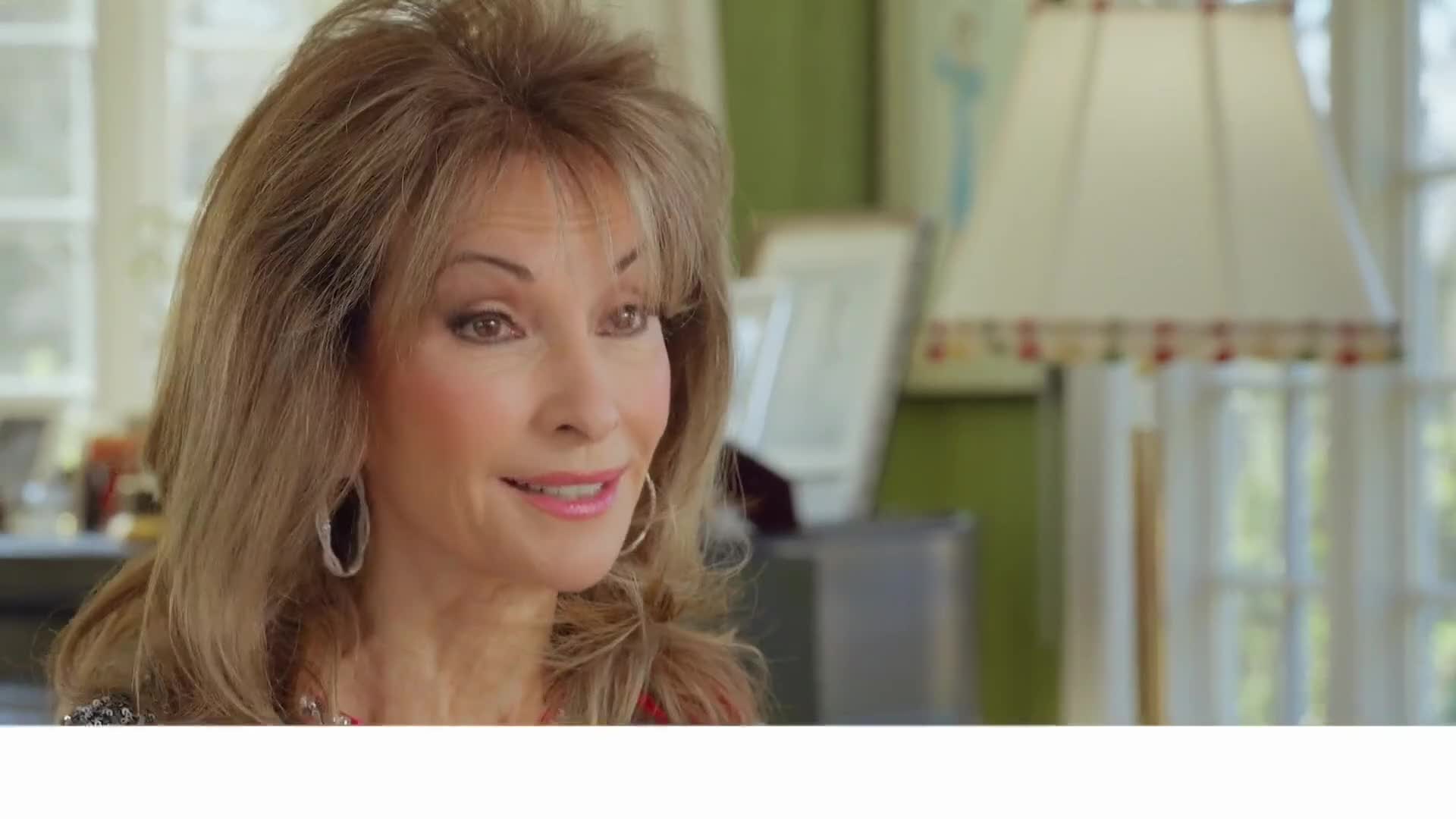 A woman with styled blonde hair smiles gently, her large hoop earrings catching the light. Behind her, a lamp with a patterned shade stands near a window.